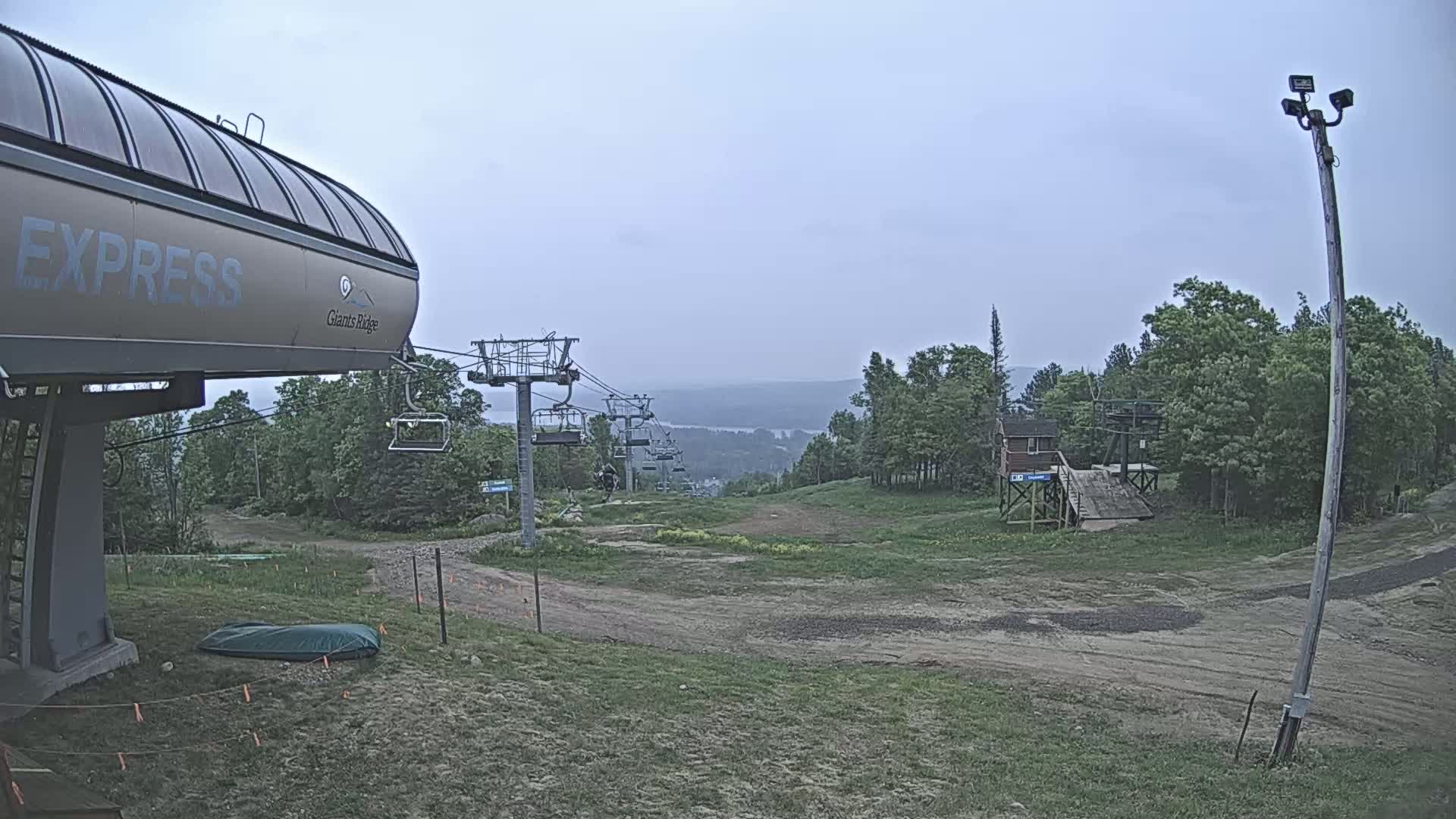 A chairlift and its base station are situated on a grassy hill overlooking a distant lake under an overcast sky.
