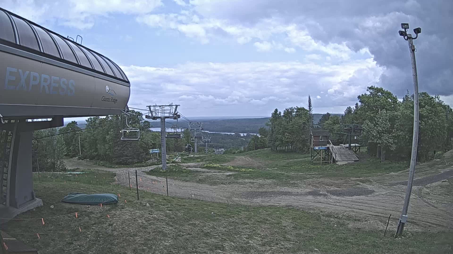 A ski lift base station and chairlift extend towards a distant view of a lake and forest under a partly cloudy sky.