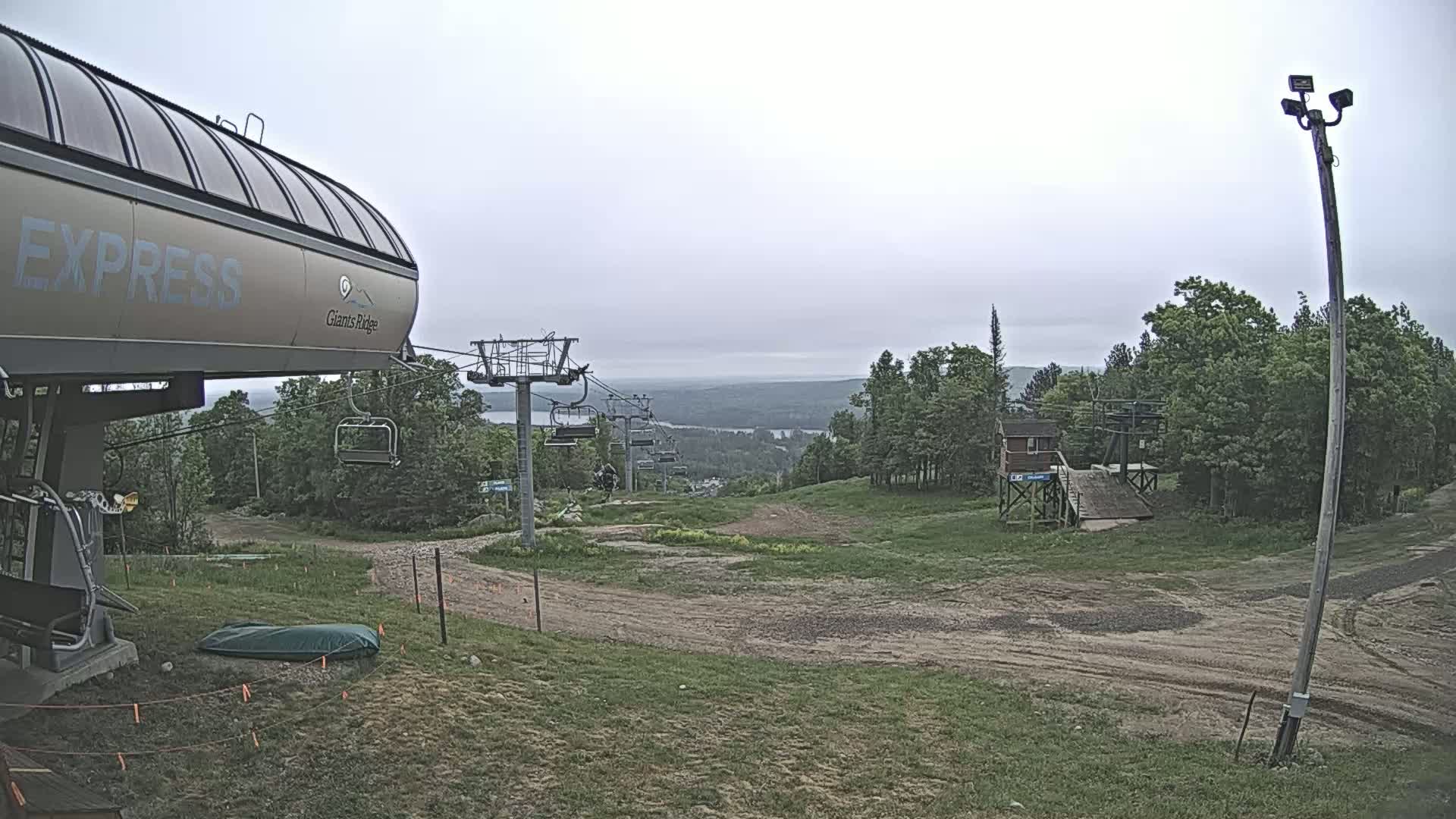 A chairlift, partially visible, sits in a grassy area overlooking a distant, hazy landscape under an overcast sky.