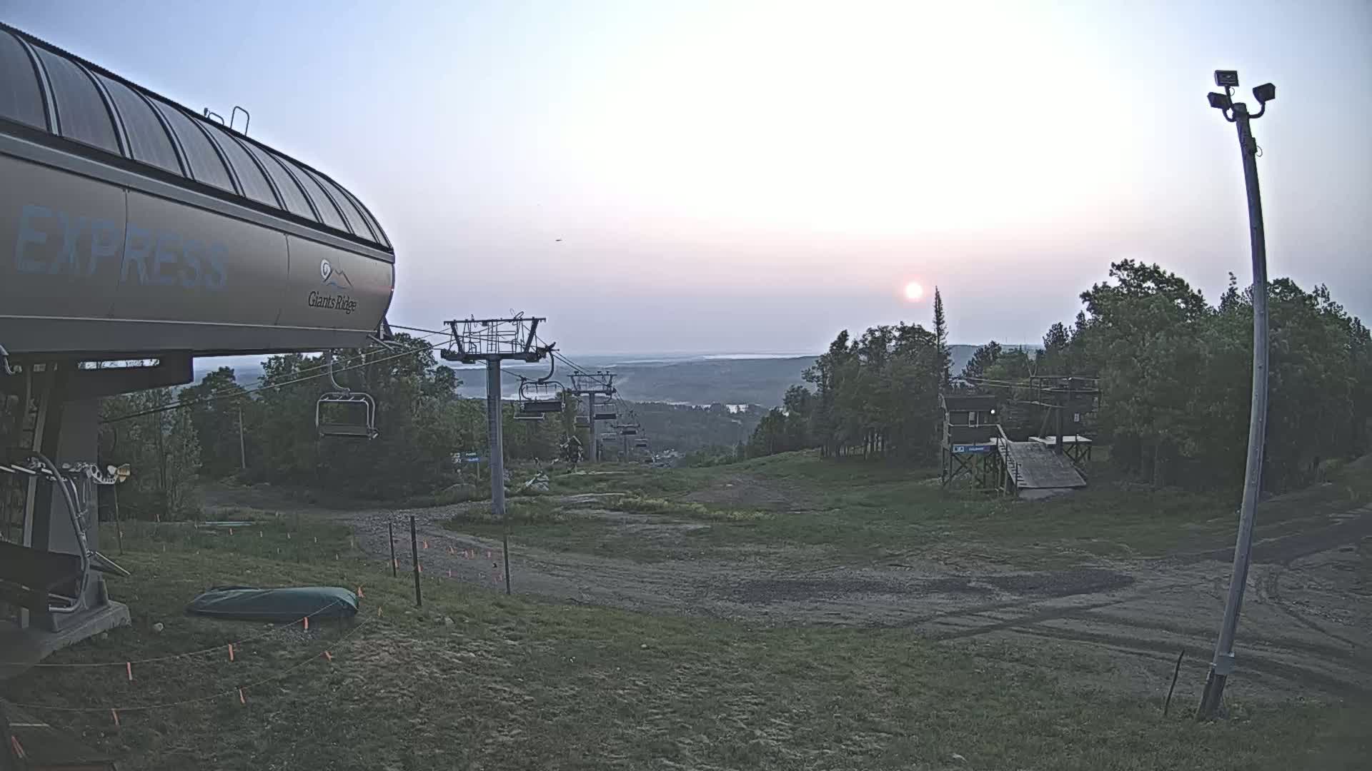 A ski lift base area at sunset, with a chairlift extending towards a distant horizon under a hazy sky.