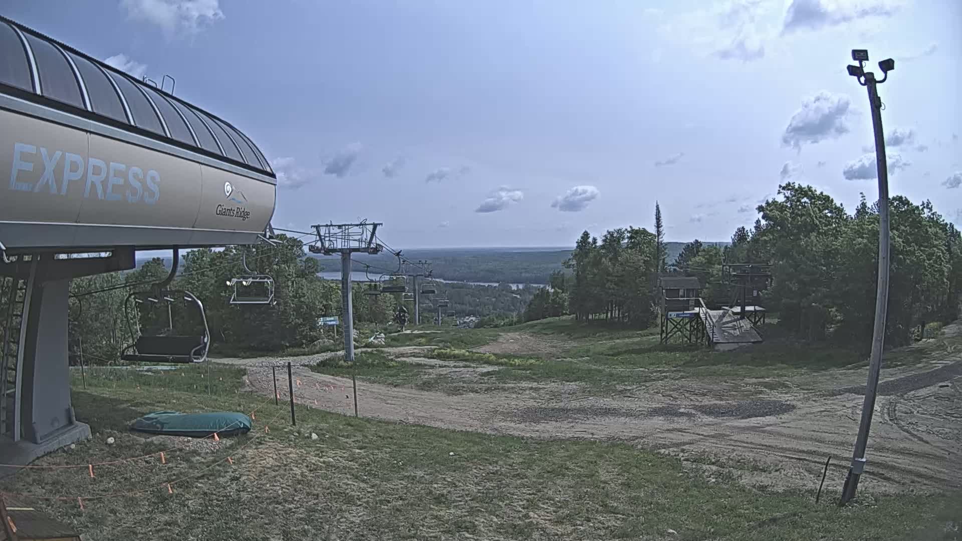 A chairlift base station and a mostly clear sky are visible, overlooking a grassy hill leading to a distant body of water under partly cloudy conditions.