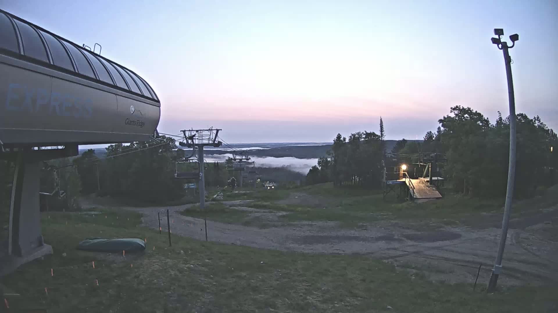 A ski lift terminal sits at the top of a hill overlooking a valley filled with low-lying fog under a twilight sky.
