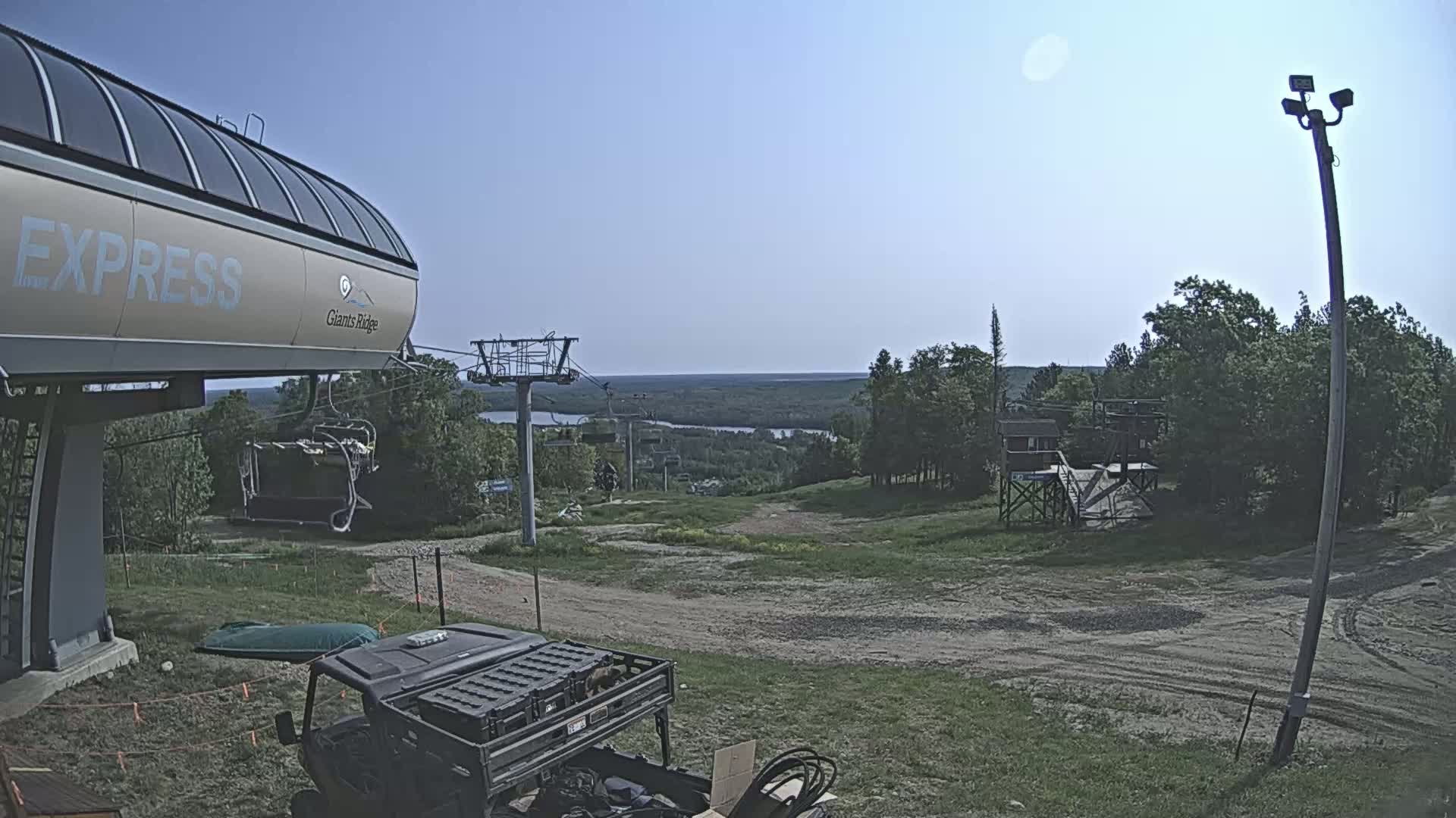 A sunny day at a ski resort shows a chairlift, a utility vehicle, and a distant view of a river and forest.