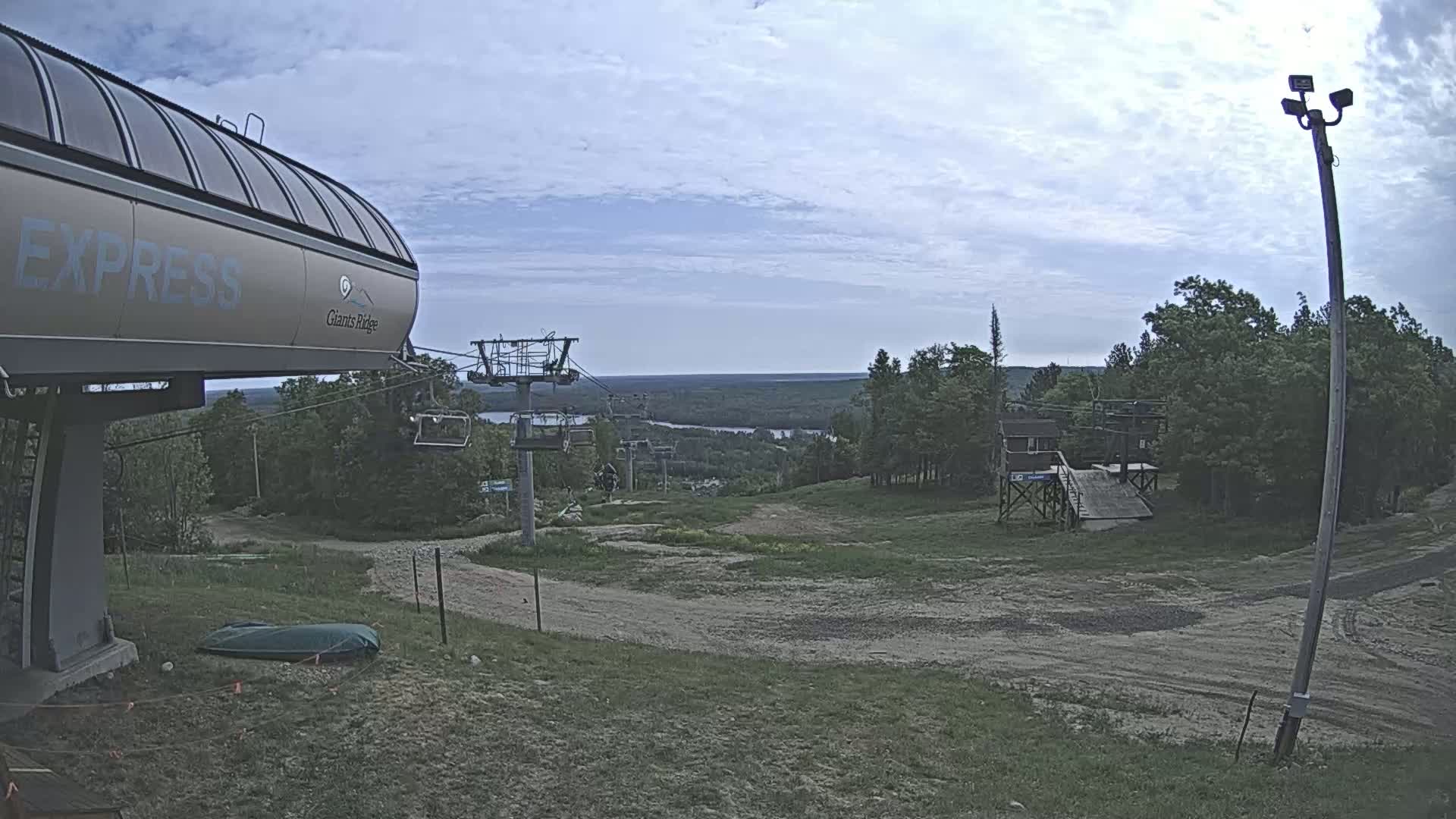 A chairlift with a large, enclosed gondola at the top overlooks a mostly clear, partly cloudy sky and a distant body of water, with a grassy hill and trees in the foreground.