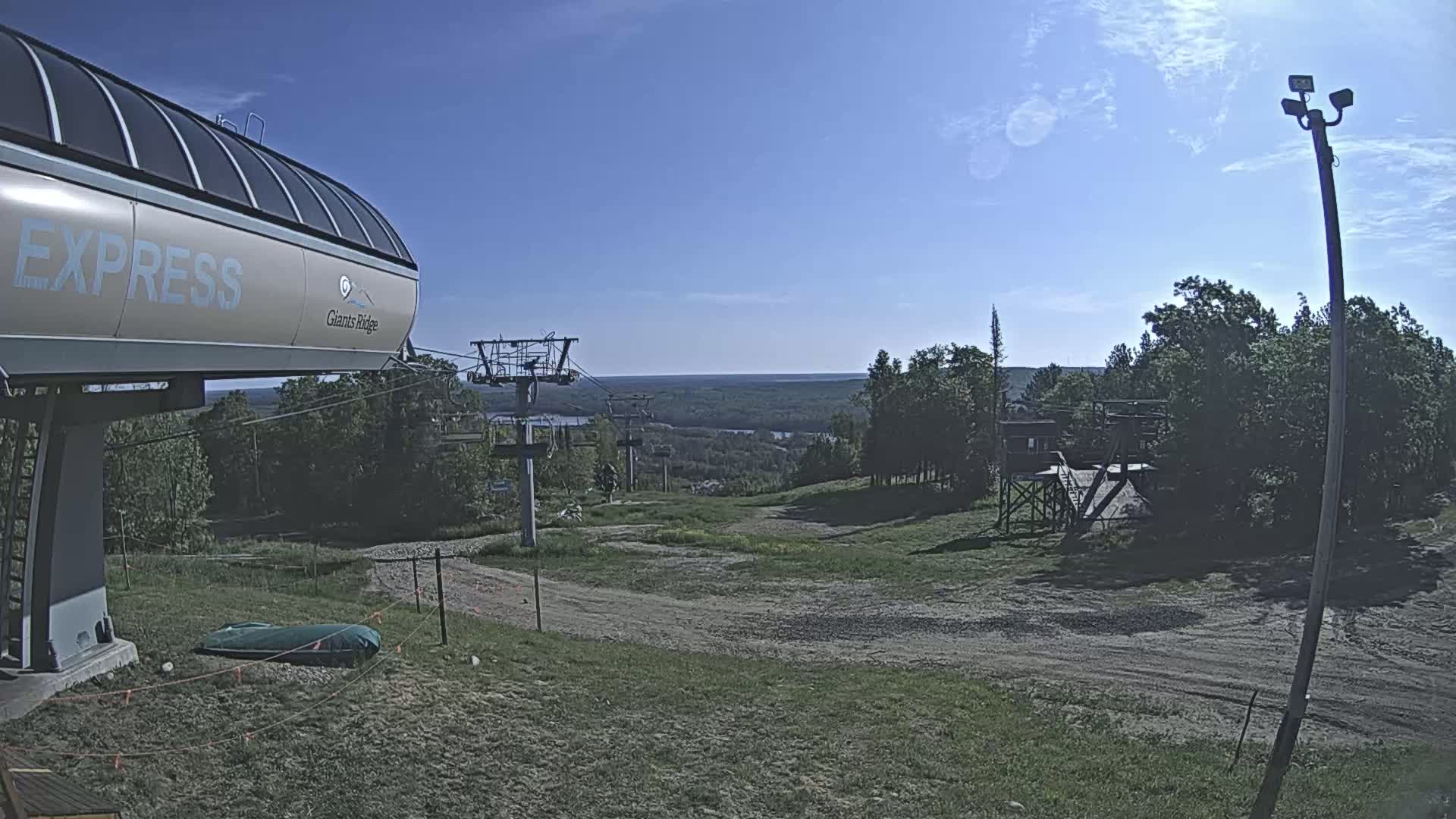 A ski lift station with a chairlift extending to a grassy hill overlooking a vast, forested landscape under a mostly clear, sunny sky.