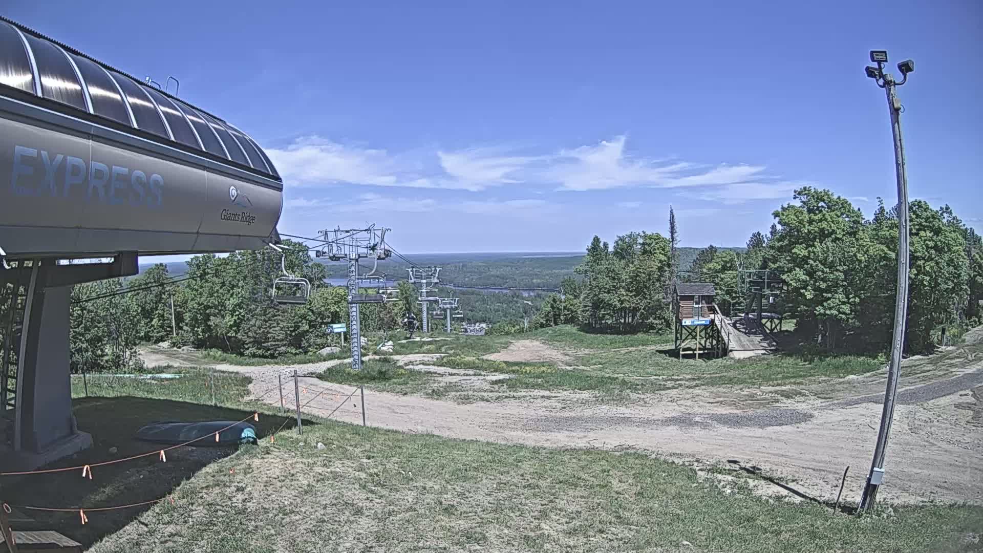A ski lift station with a chairlift extending to a grassy hill overlooking a vast, forested landscape under a mostly clear, sunny sky.