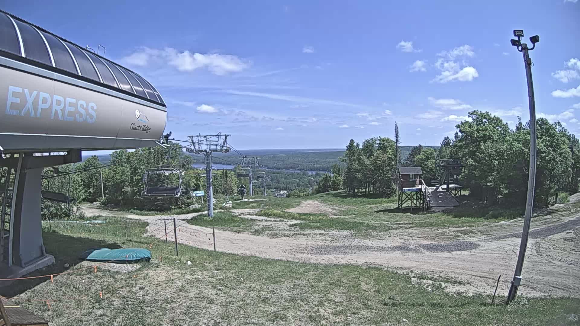 A ski lift base area with a chairlift and a view of a lake and forest under a partly cloudy blue sky.