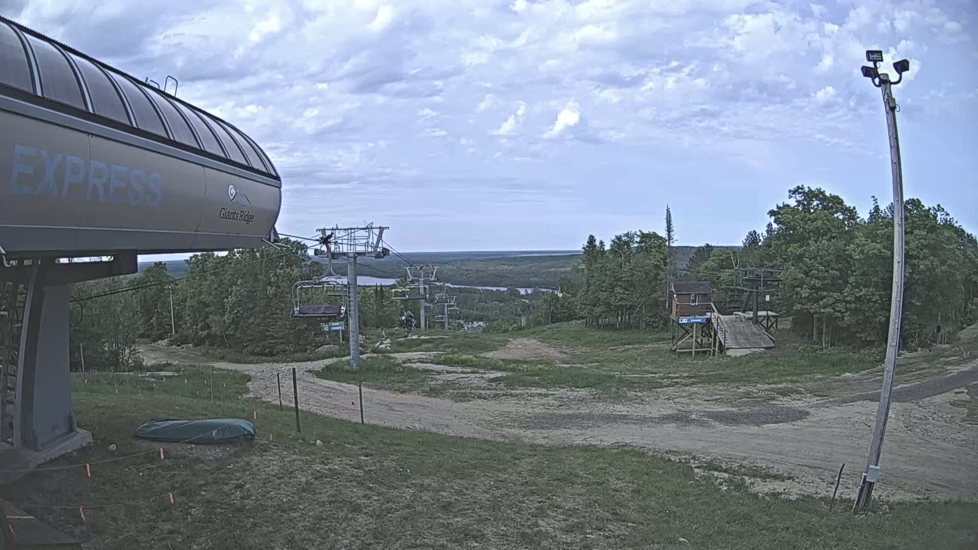 A ski lift base area with a chairlift and a view of a lake and forest under a partly cloudy blue sky.