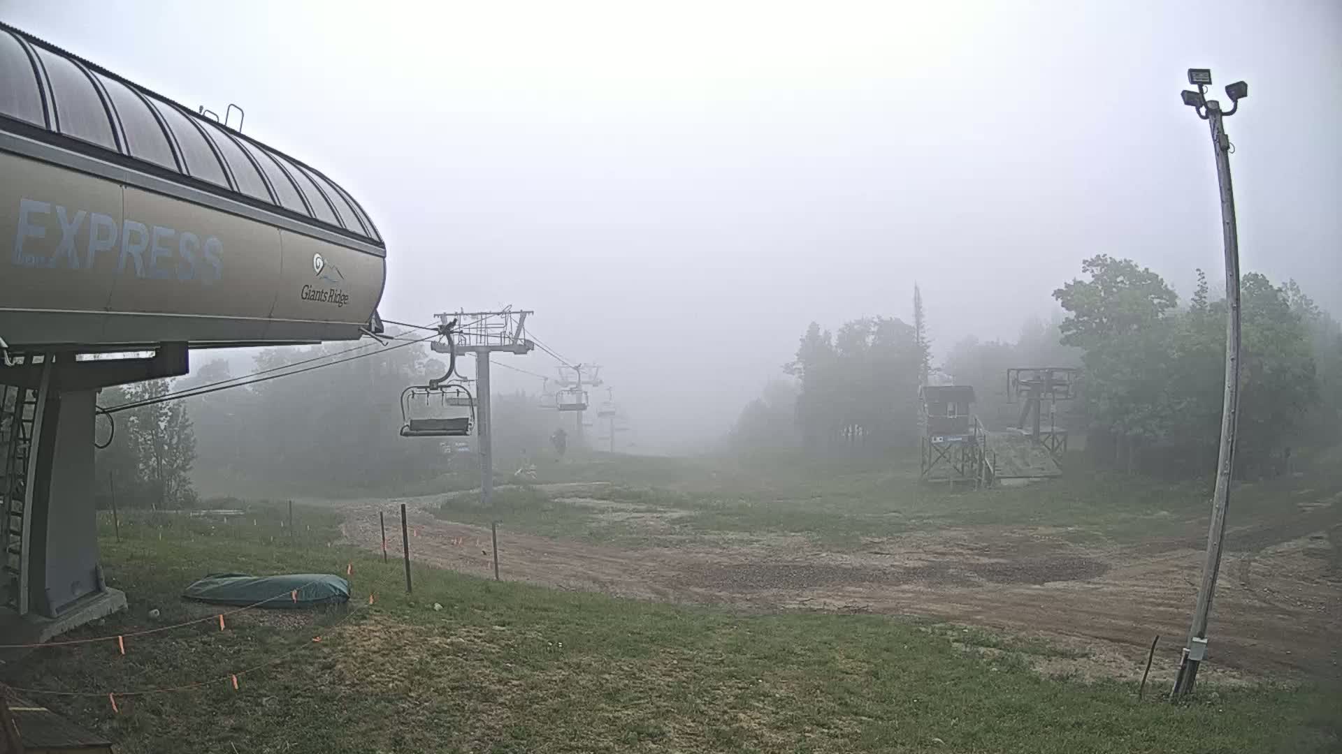 A ski lift base station and chairlifts are partially visible through thick fog on a grassy area with a dirt path.