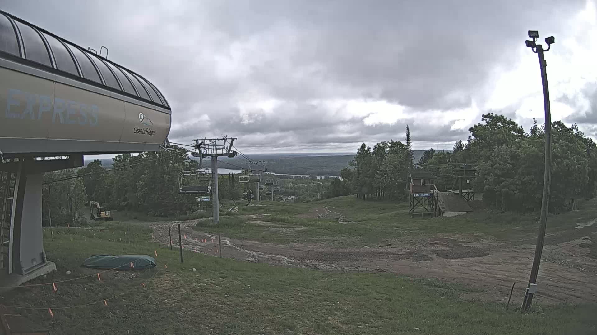 A chairlift, a small building, and a grassy area with a dirt path are visible under a cloudy sky, overlooking a distant body of water and forested hills.