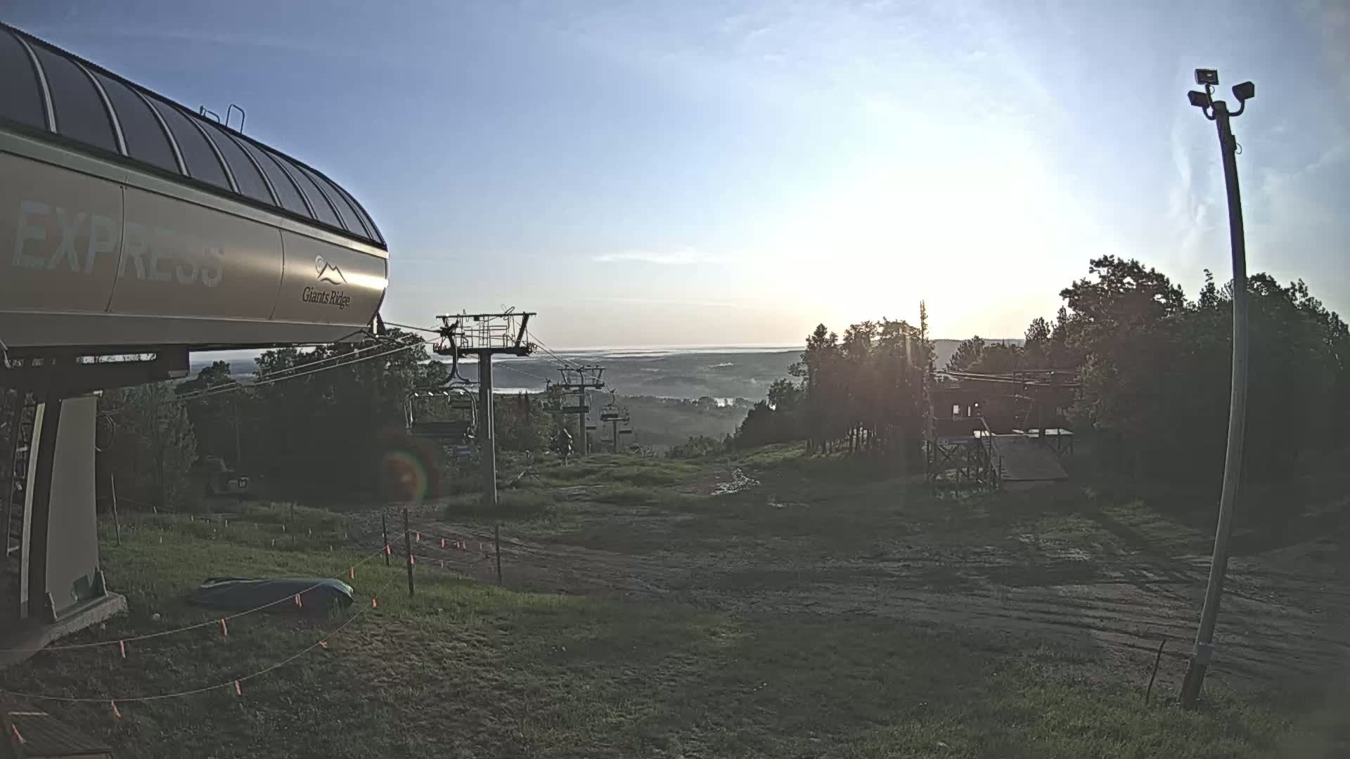 A chairlift base station sits at the top of a grassy hill overlooking a distant body of water under a clear, sunrise sky.