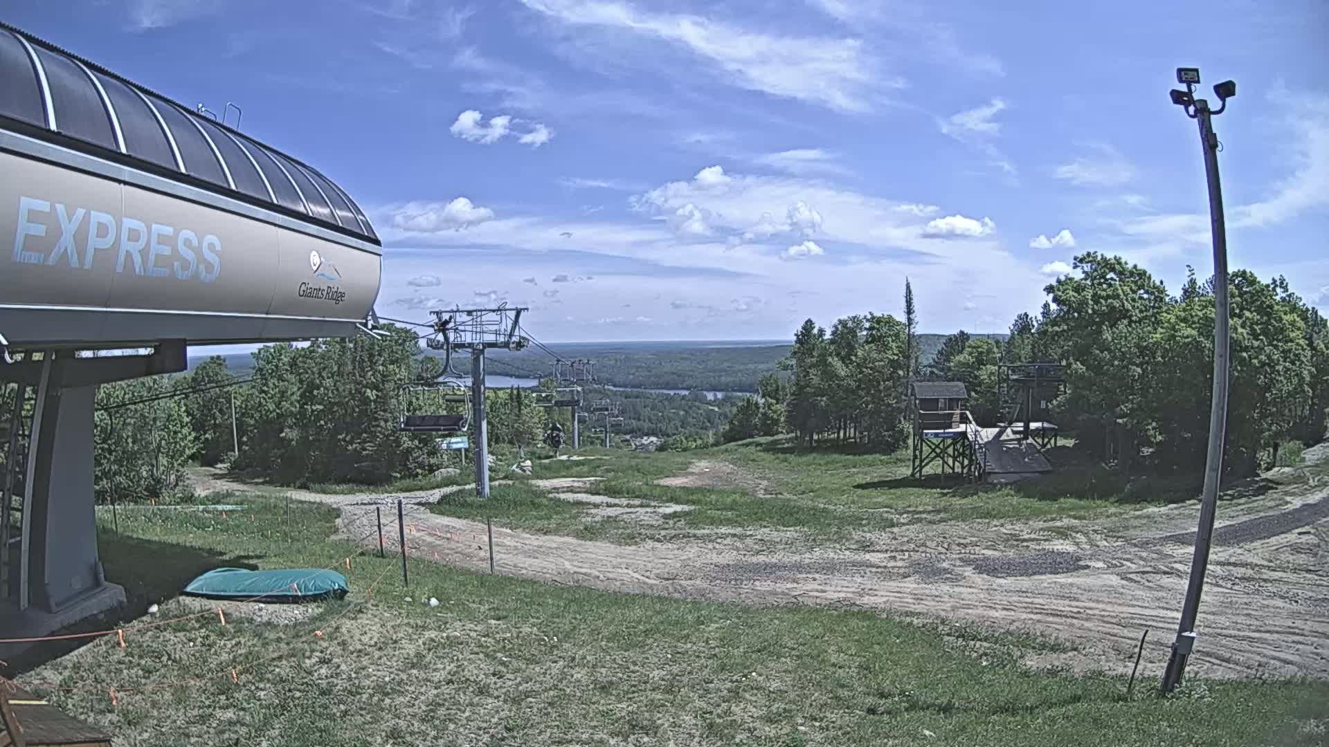 A ski lift base station on a partly cloudy day overlooks a grassy hill and a distant body of water.
