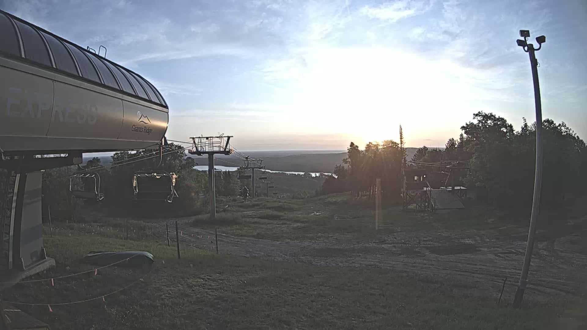 A chairlift at sunset overlooks a calm lake and forested hills under a mostly clear sky.