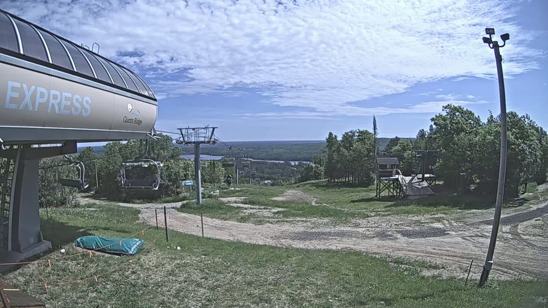 A ski lift base station with a chairlift ascending a grassy hill towards a distant river under a partly cloudy sky.