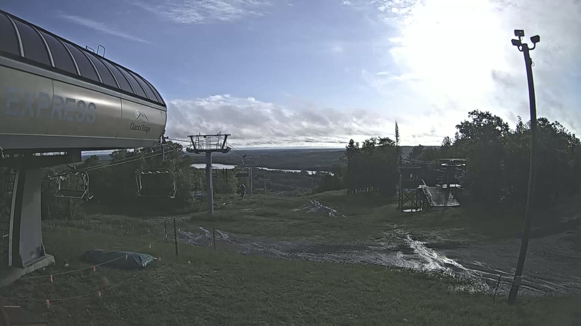 A chairlift at a ski resort is visible under a partly cloudy sky, with a distant view of a lake and forested landscape.