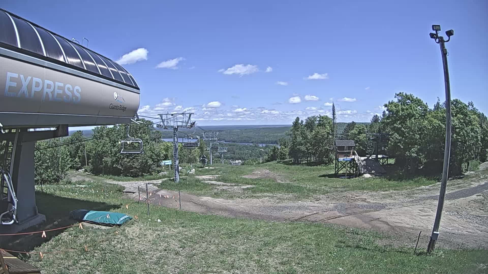 A ski lift base area with a chairlift extending to a distant body of water under a mostly sunny sky.