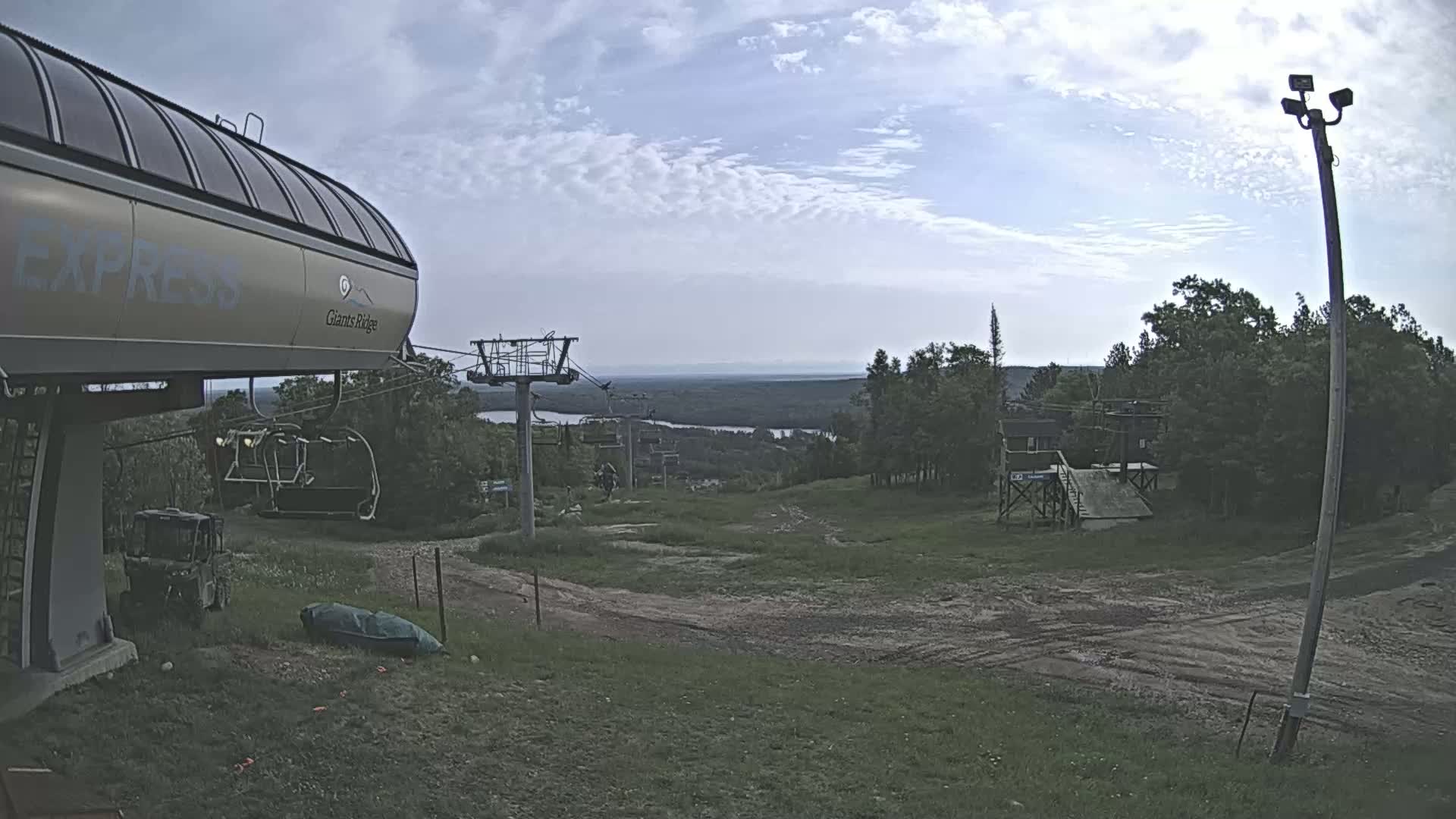 A ski lift base station with overcast skies overlooks a grassy area leading to a distant body of water and forested hills.