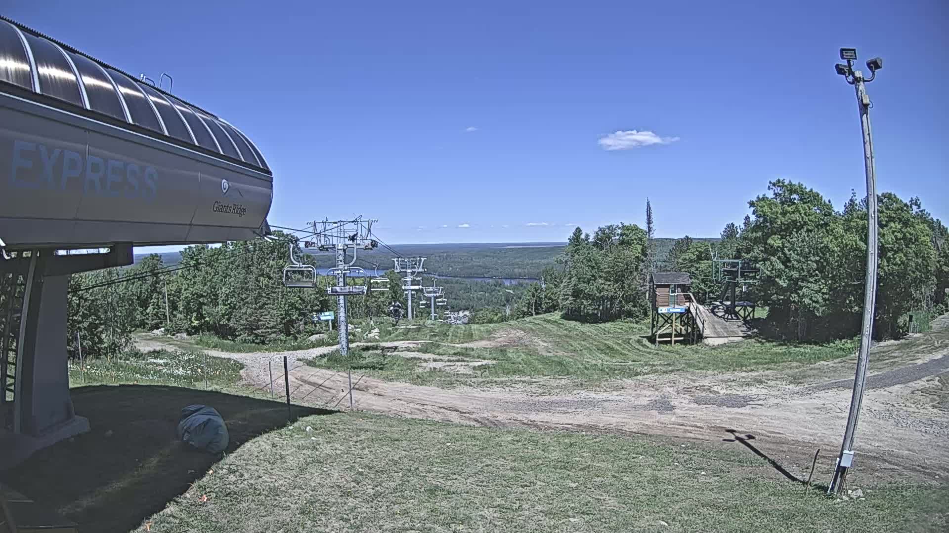 A chairlift base station and a view of a grassy ski hill extending to a lake and forested landscape under a mostly clear blue sky.