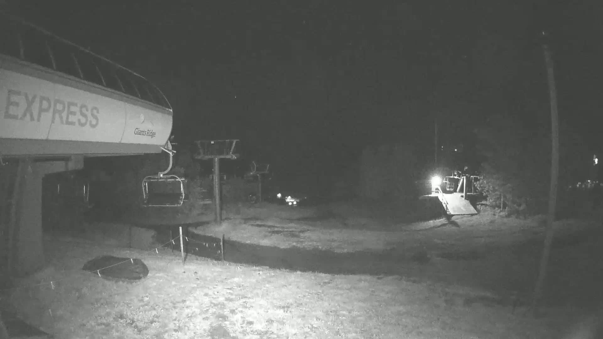 A nighttime, low-light grayscale image shows a chairlift and its base station, with a small loading ramp visible in the background.