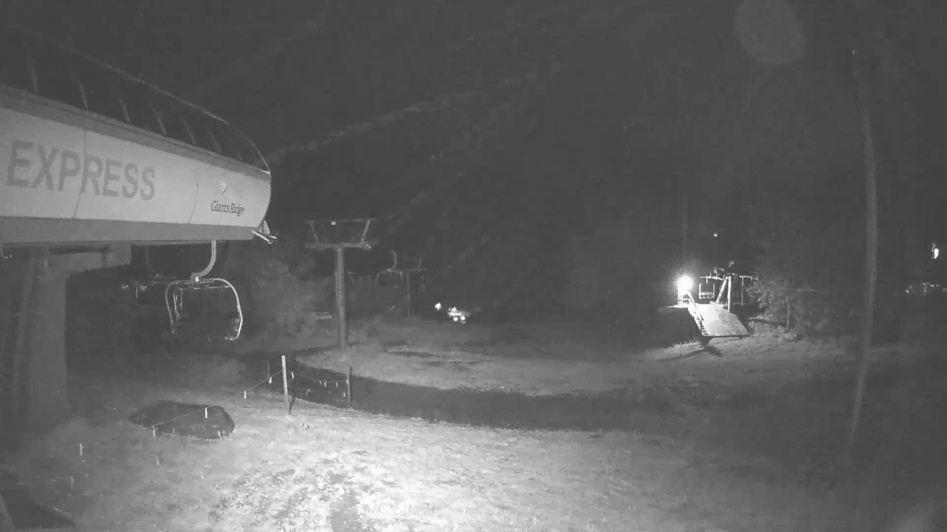 A nighttime, snow-covered ski lift base area with a chairlift partially visible.