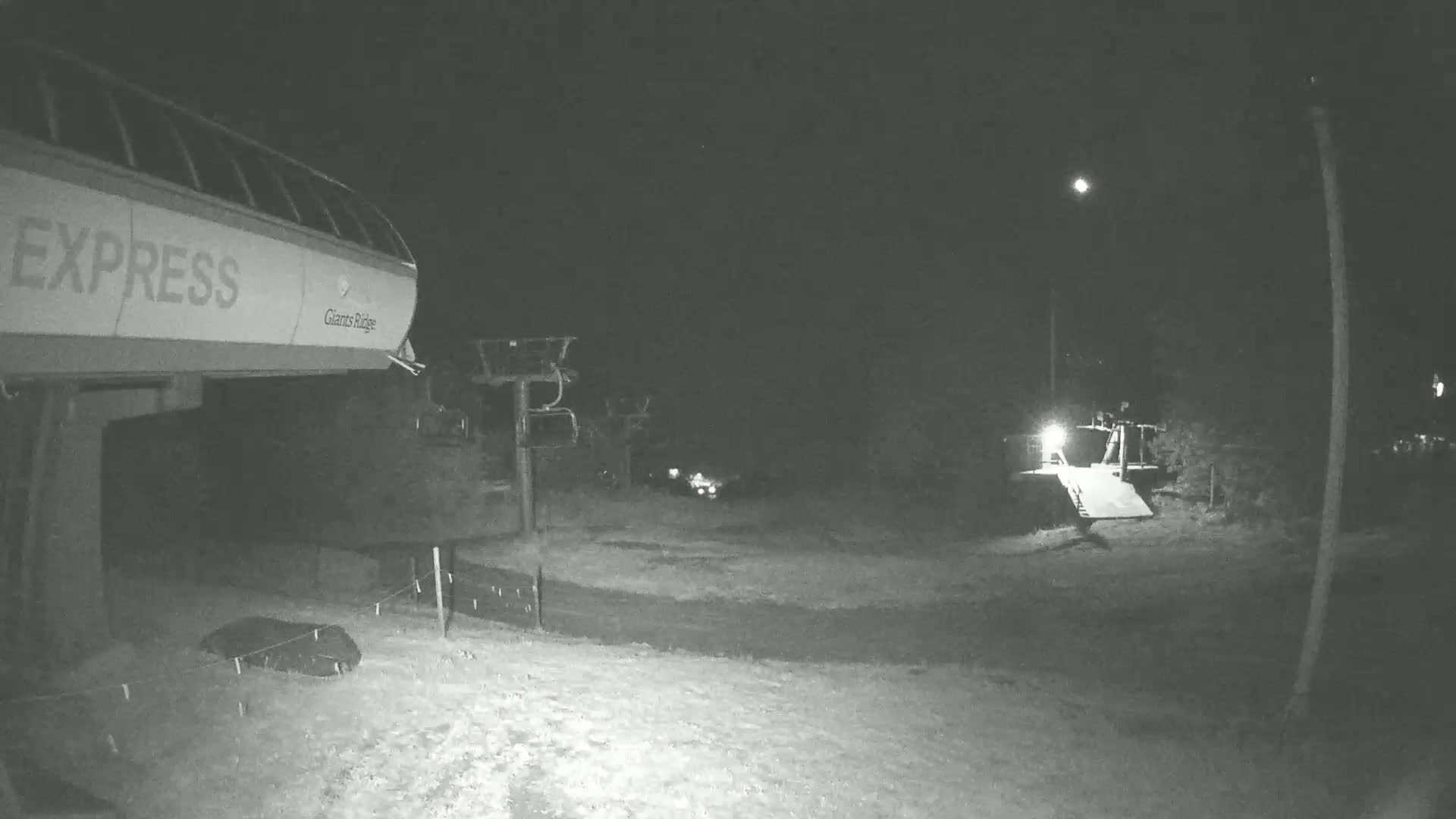 A nighttime, snow-covered ski lift base area with a chairlift in the foreground and other lifts and structures visible in the distance under a moonlit sky.