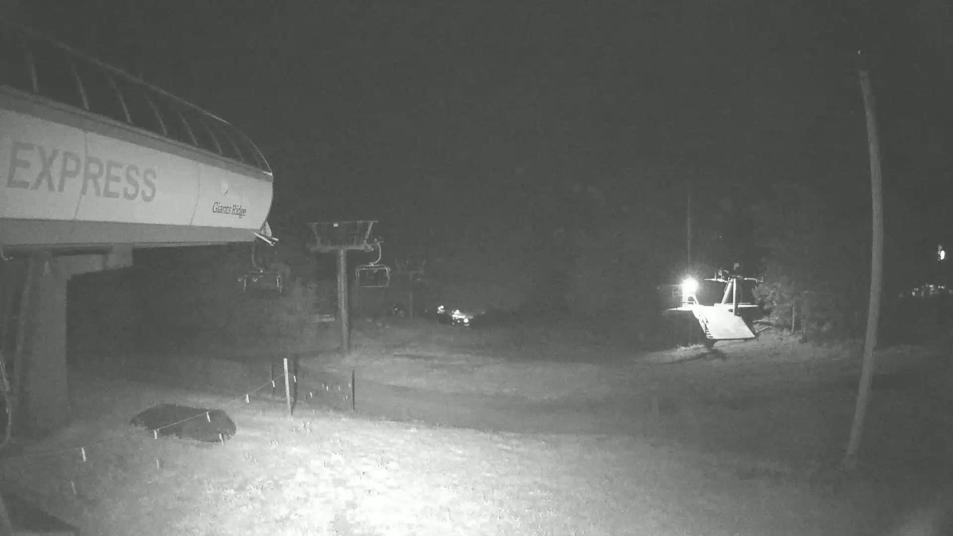 A nighttime, snow-covered ski lift base area with a chairlift in the foreground and other lifts and structures visible in the distance under a moonlit sky.