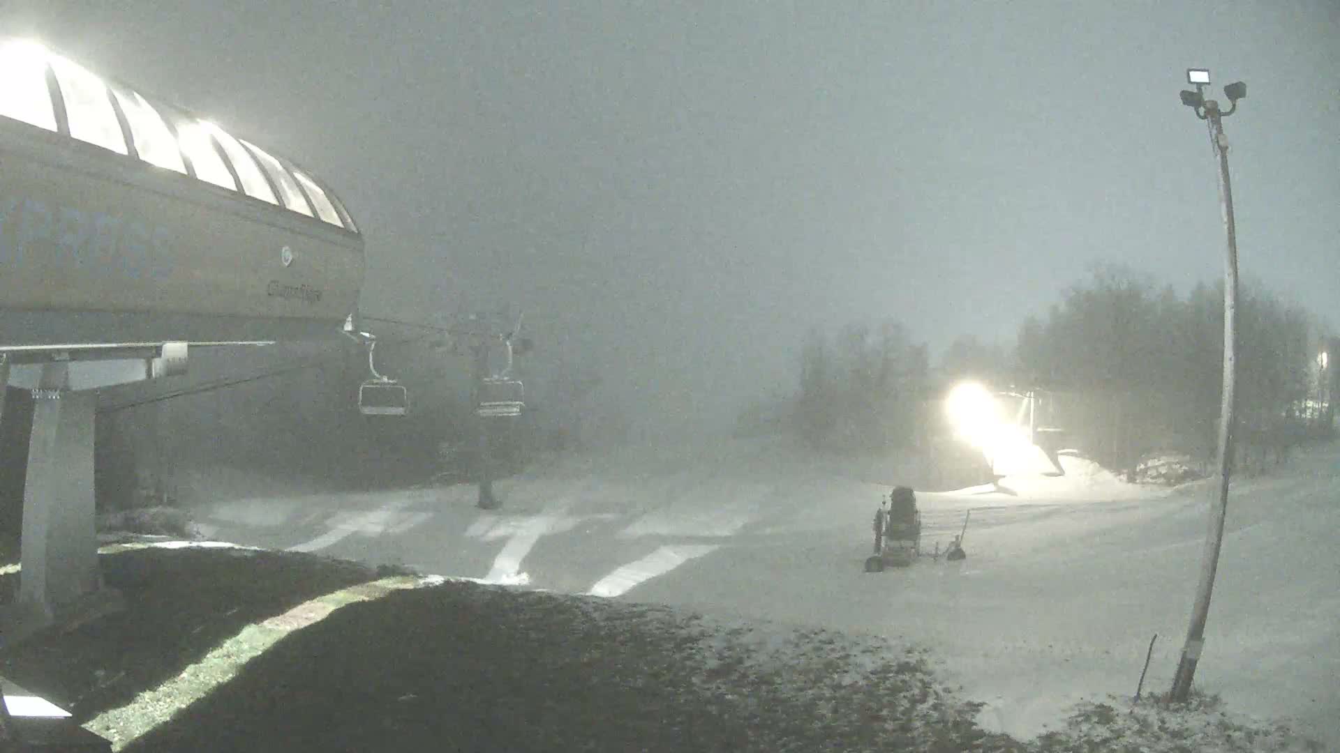 An illuminated ski lift station and a bright snow machine operate on a snow-covered ski slope at night, with the entire scene enveloped in dense fog.