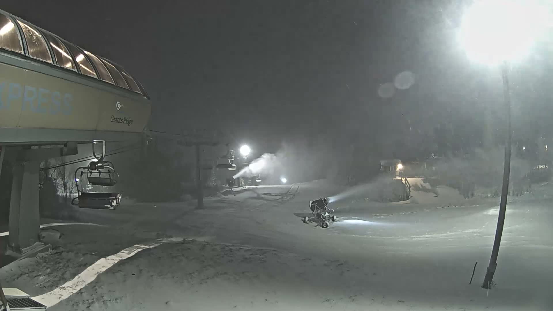 The image depicts a snowy night at a ski resort, where a ski lift stands in the foreground while snow guns and a snow groomer are actively producing snow on the brightly lit slopes.
