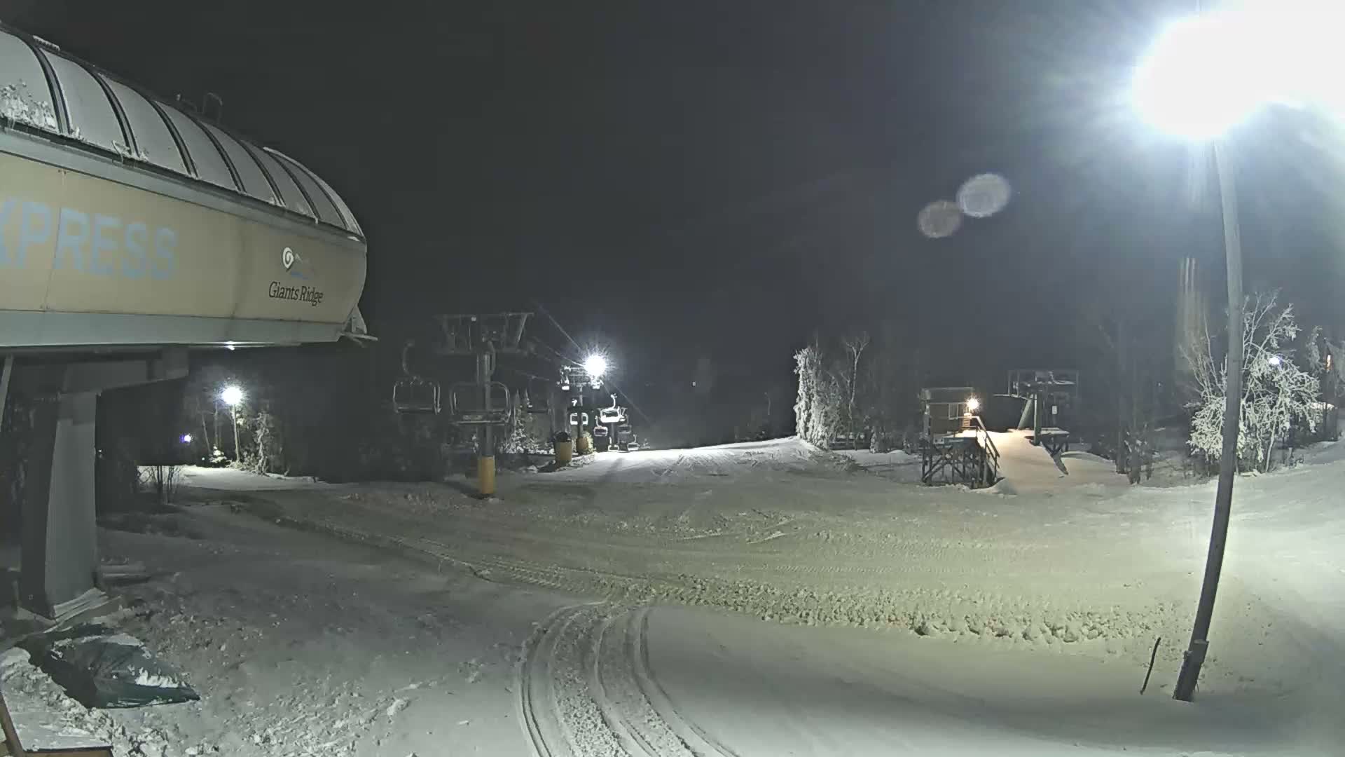 A brightly lit ski resort is seen at night under a clear sky, with snow covering the ground, trees, and ski lifts, and ski tracks visible on the slopes.