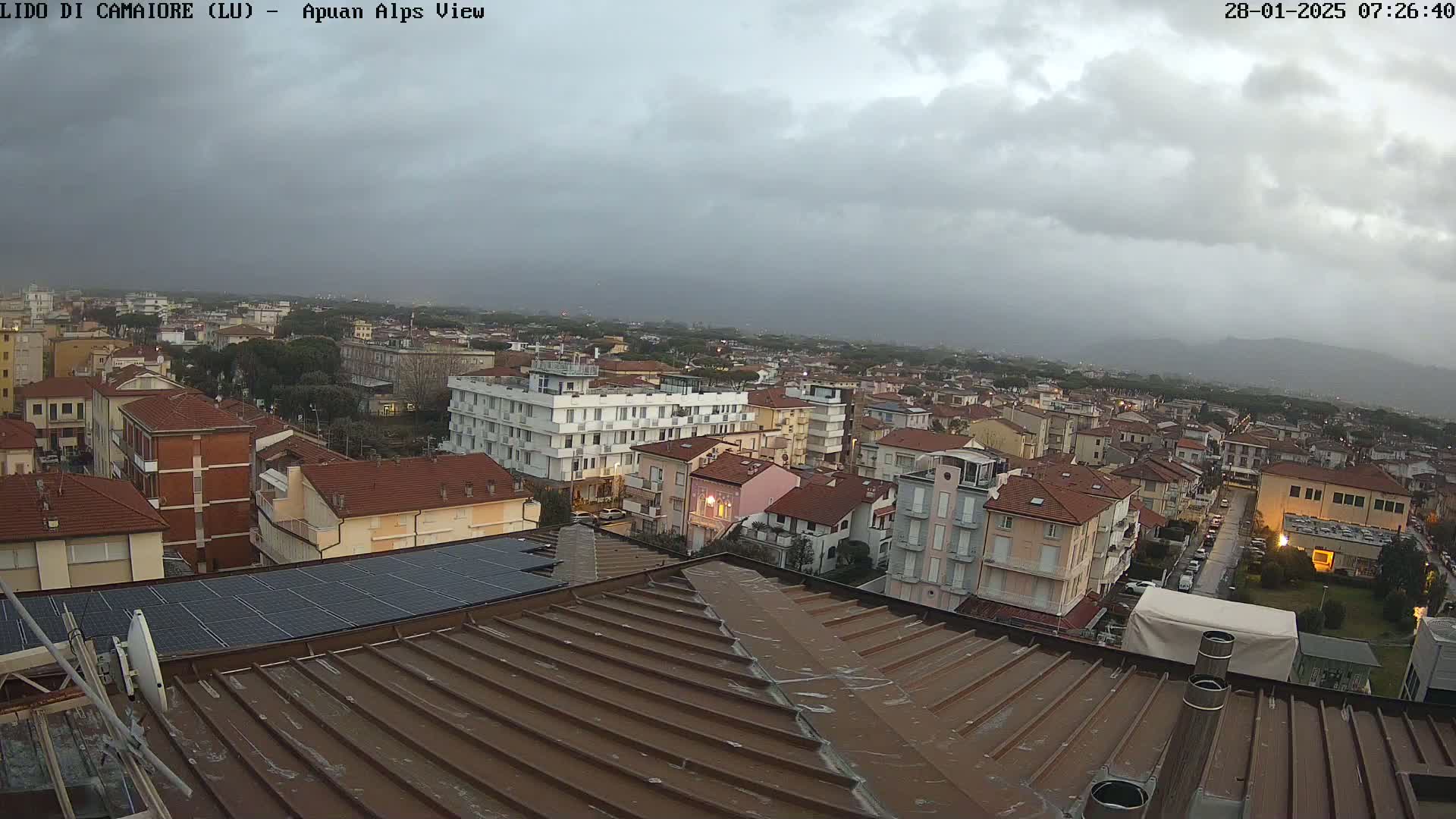 A rooftop view of a town under an overcast sky, showing many low-rise buildings extending to distant mountains.