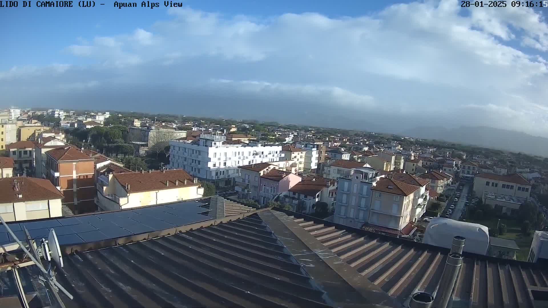A partly cloudy sky overlooks a sprawling town of low-rise buildings with reddish-brown roofs, seen from a rooftop with solar panels.