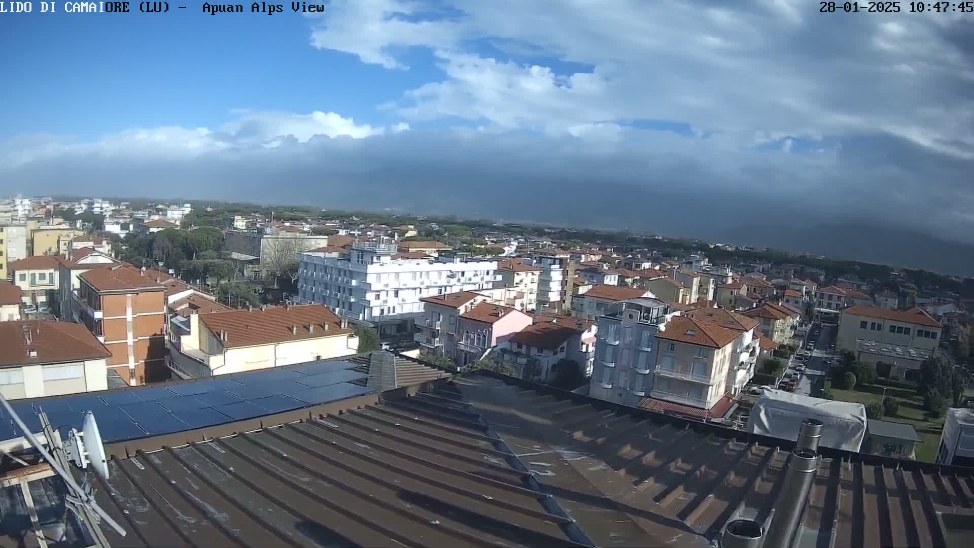A mostly sunny day reveals a wide view of a town's rooftops and buildings, stretching to a distant horizon under a partly cloudy sky.
