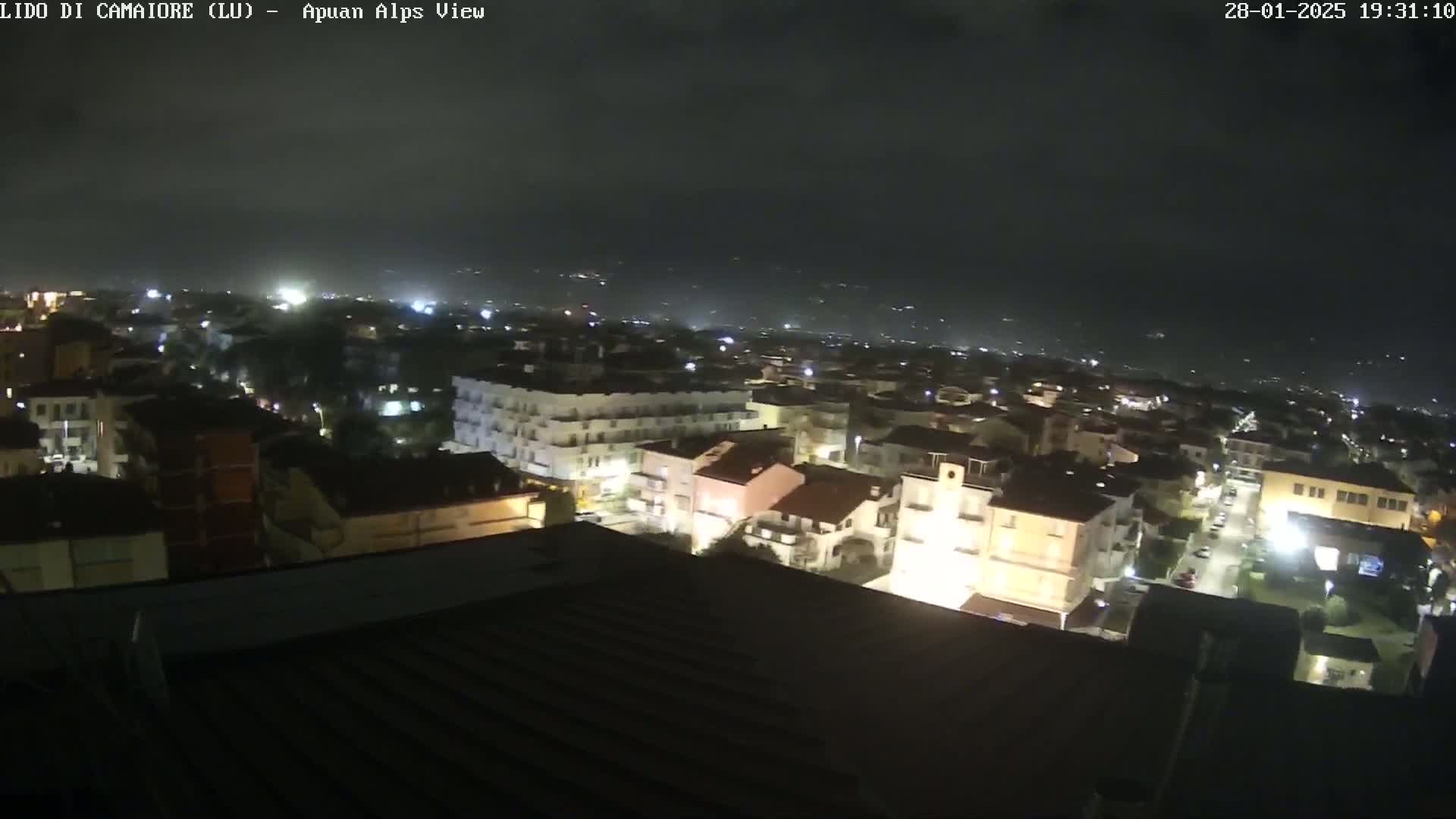 A nighttime aerial view of a town shows many buildings illuminated under a dark, cloudy sky.
