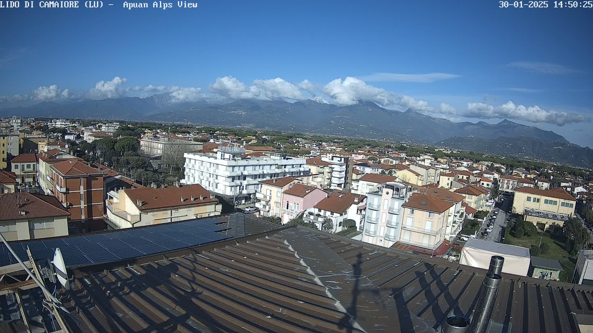 A mostly sunny day reveals a panoramic view of a town nestled beneath a mountain range, seen from a rooftop with solar panels.