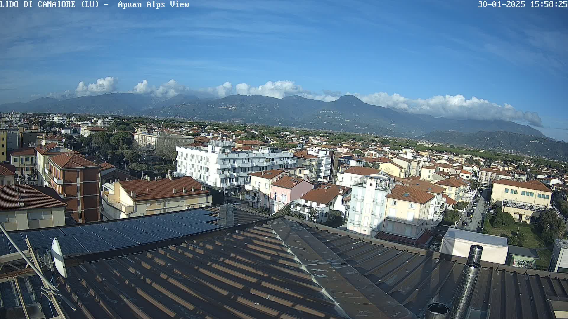A rooftop view of a town under a mostly clear blue sky with some clouds, overlooking mountains in the distance.
