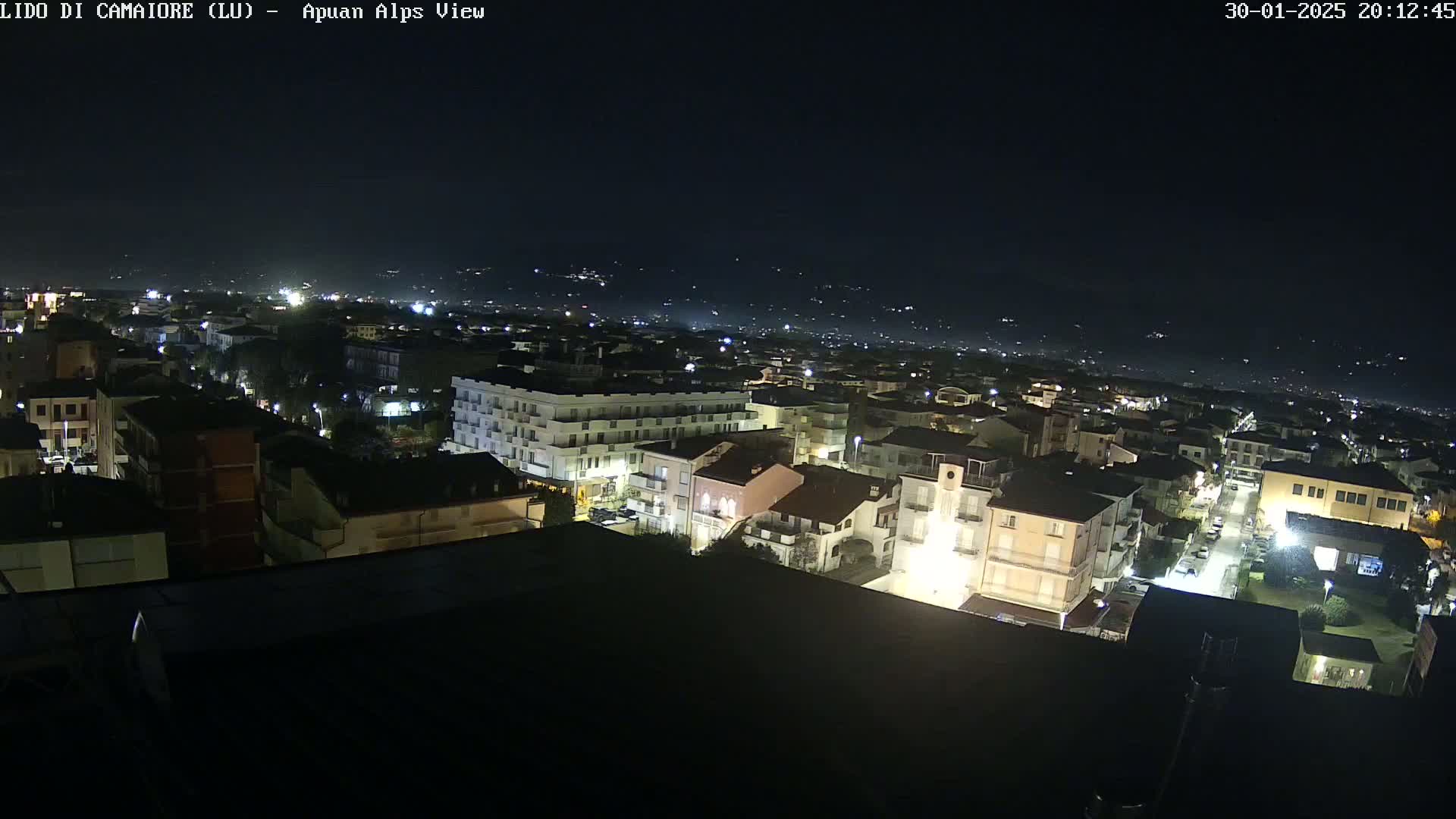 A nighttime, high-angle view of a town with illuminated buildings and streets, set against a dark sky and distant hills.