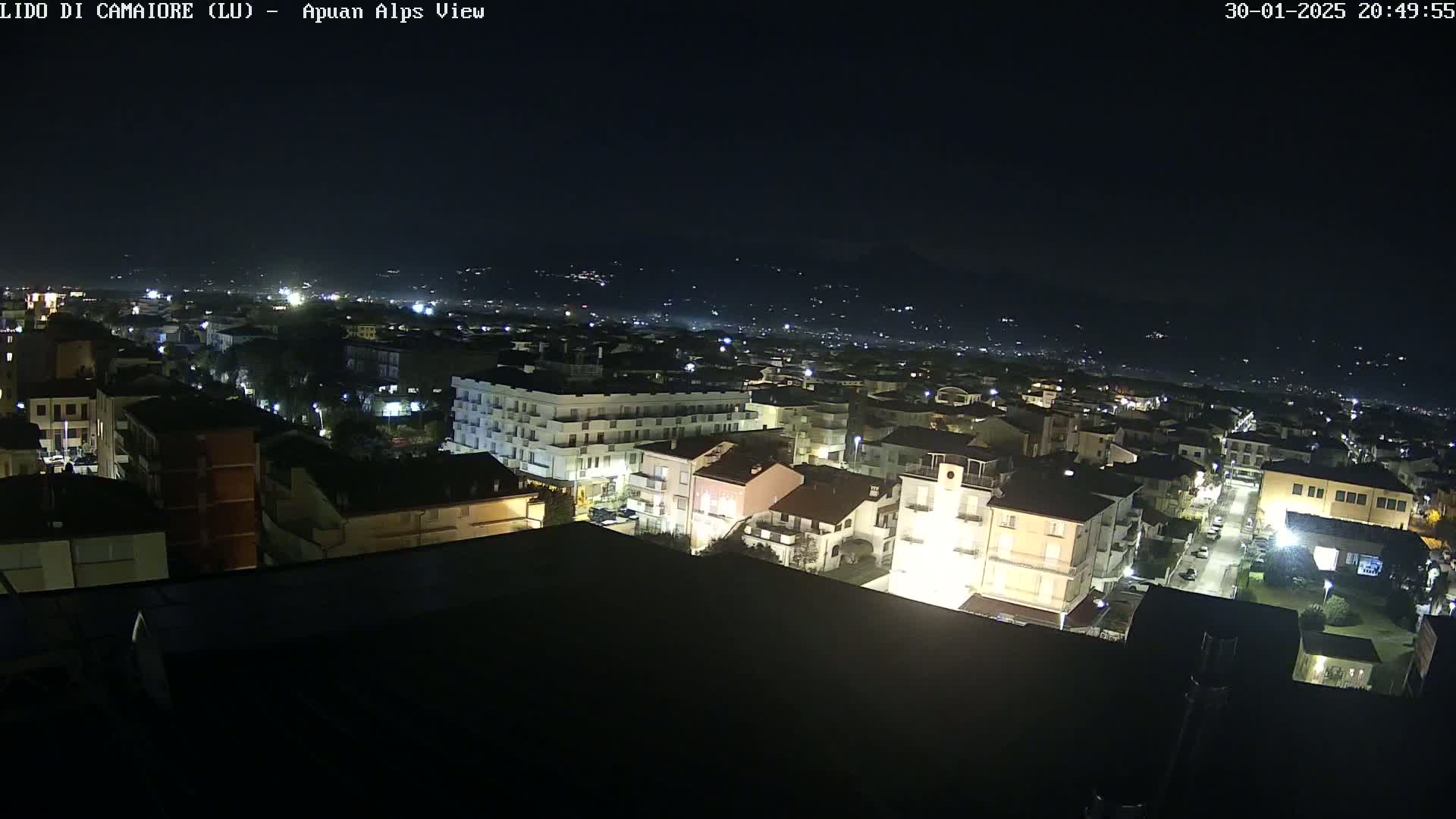 A nighttime aerial view of a town illuminated by streetlights, with a mountain range faintly visible in the distance under a clear, dark sky.
