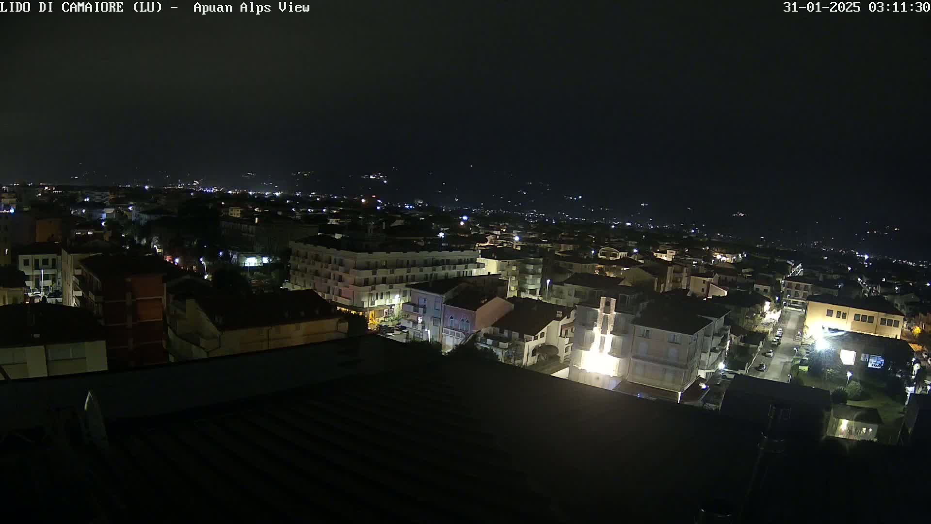 A nighttime aerial view of a town shows many buildings illuminated, with darker hills visible in the distance under a clear, dark sky.