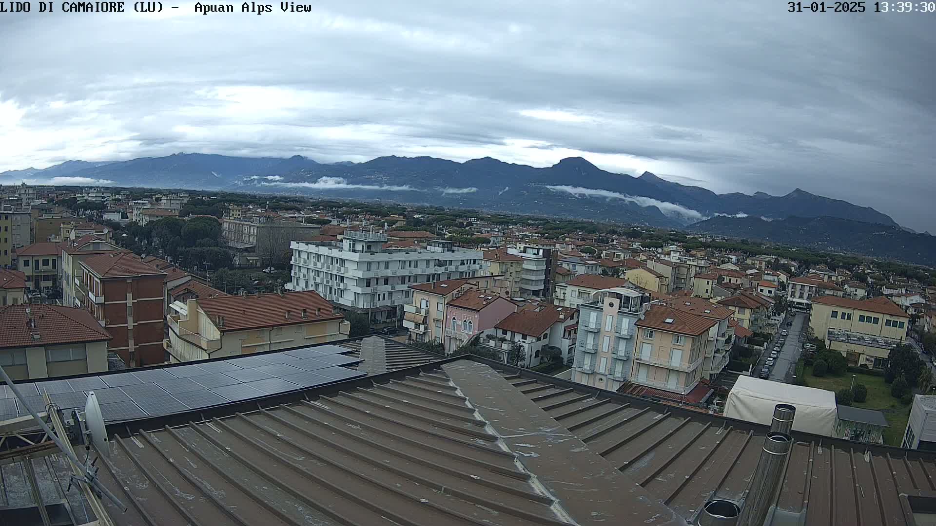 A view from a rooftop shows a town with low-rise buildings under a cloudy sky, with mountains in the background.