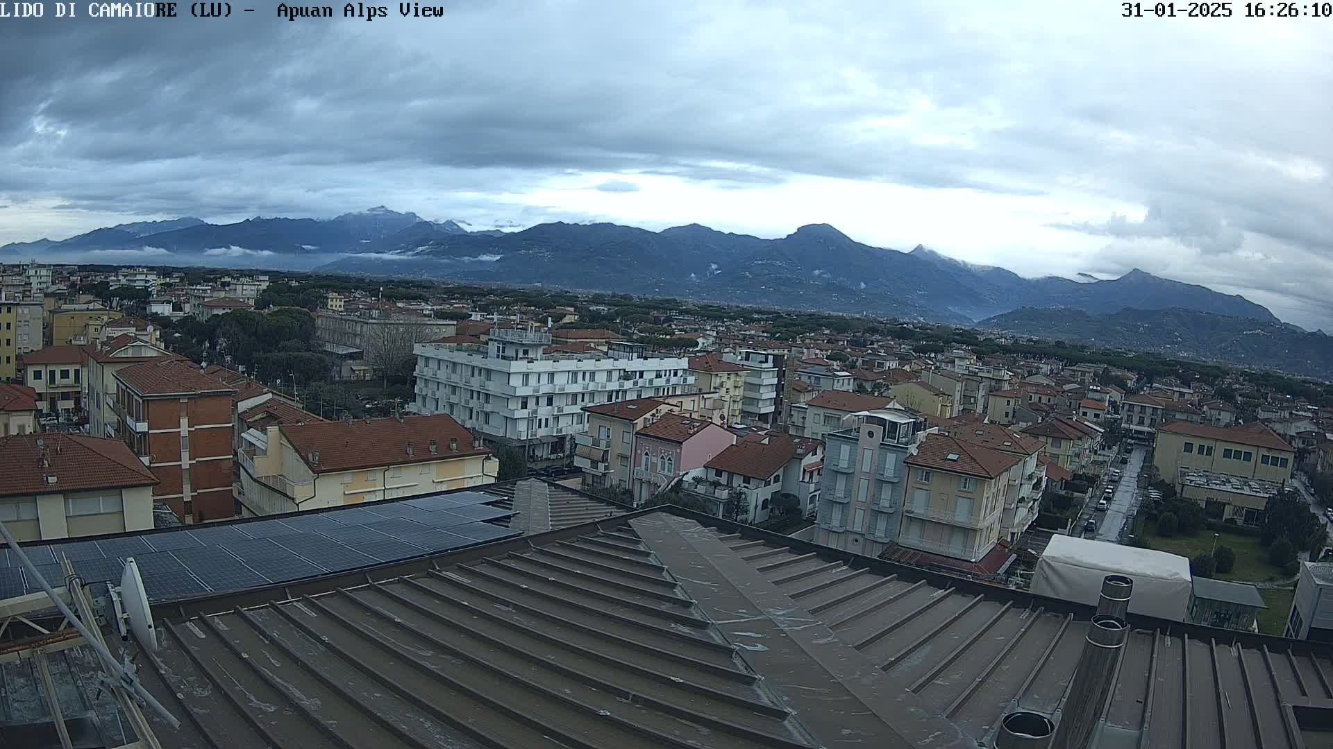 A cloudy day reveals a view of a town nestled at the foot of a mountain range, as seen from a rooftop with solar panels.
