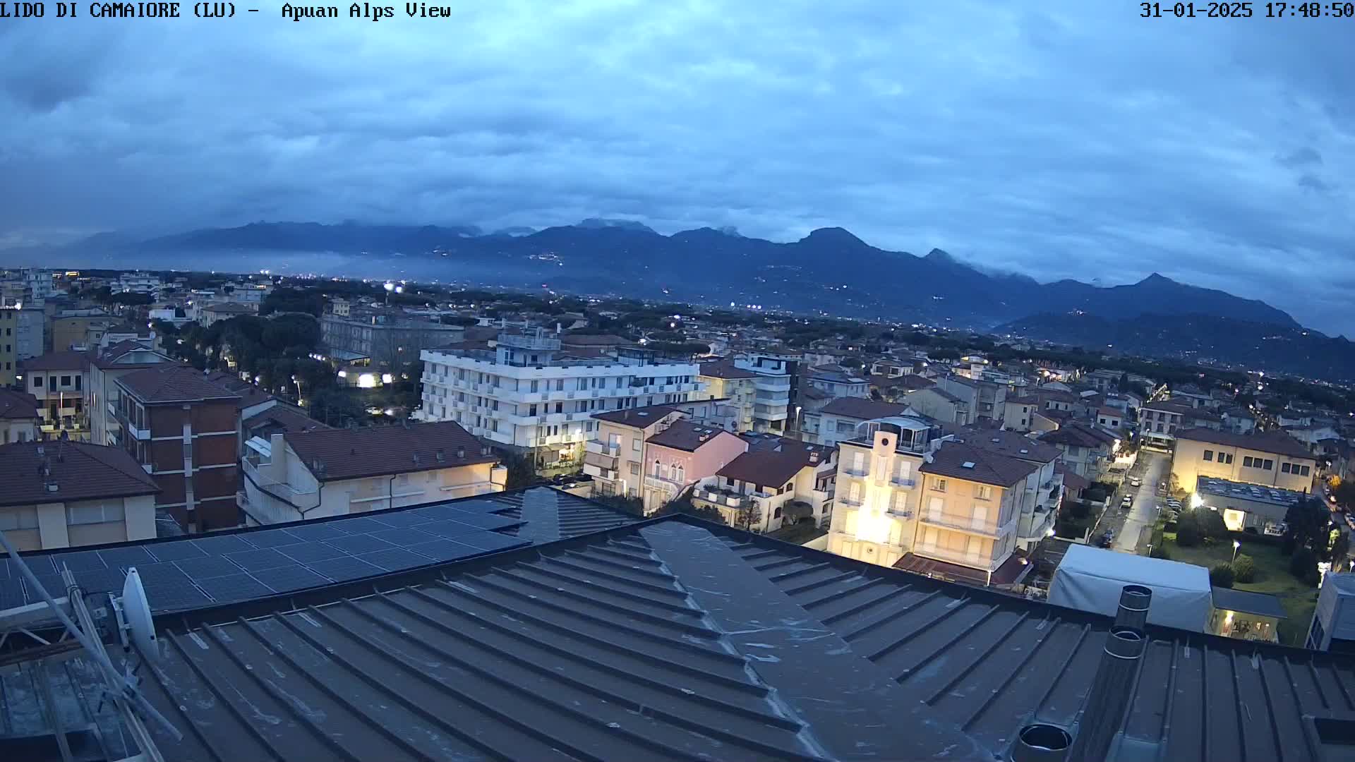 A nighttime view of a coastal town, under a mostly cloudy sky, with mountains in the background.