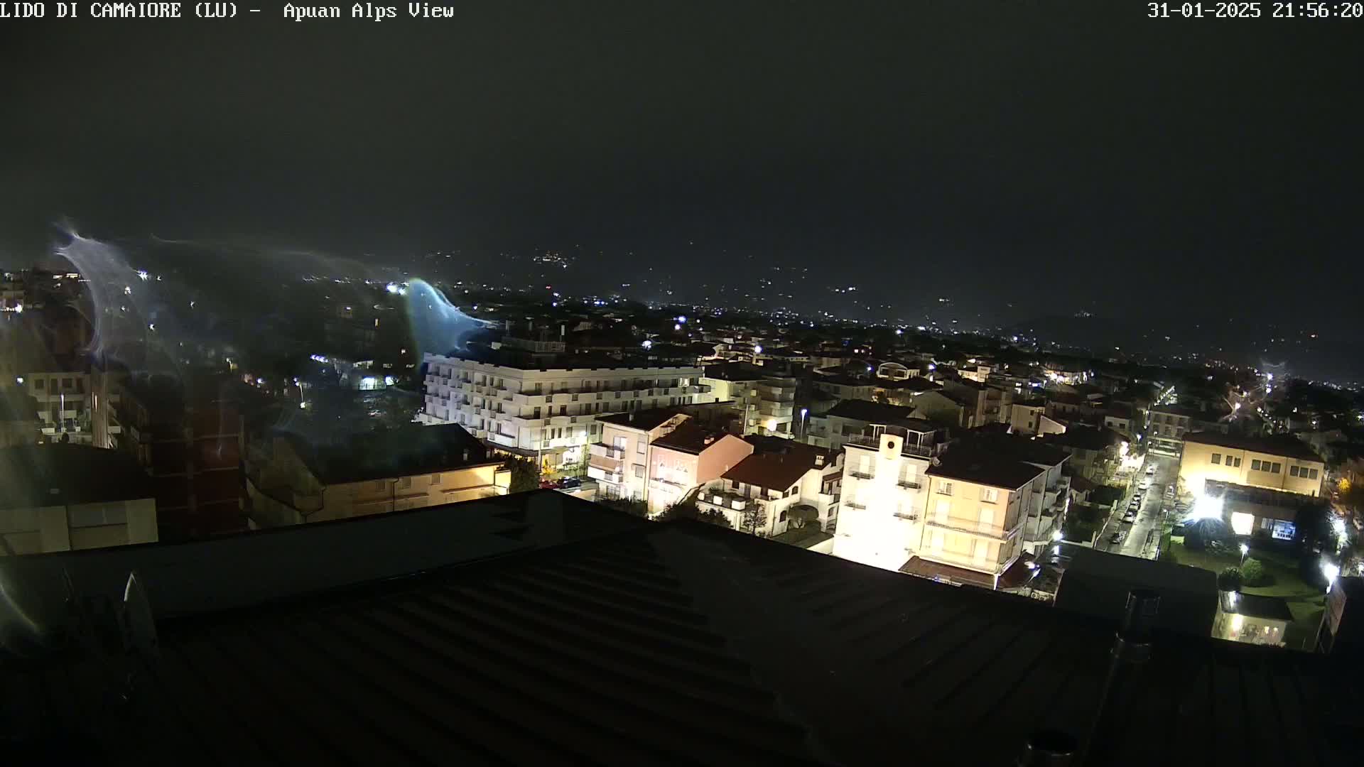 A nighttime aerial view of a town illuminated by streetlights, with faint, wispy clouds or atmospheric phenomena in the sky.