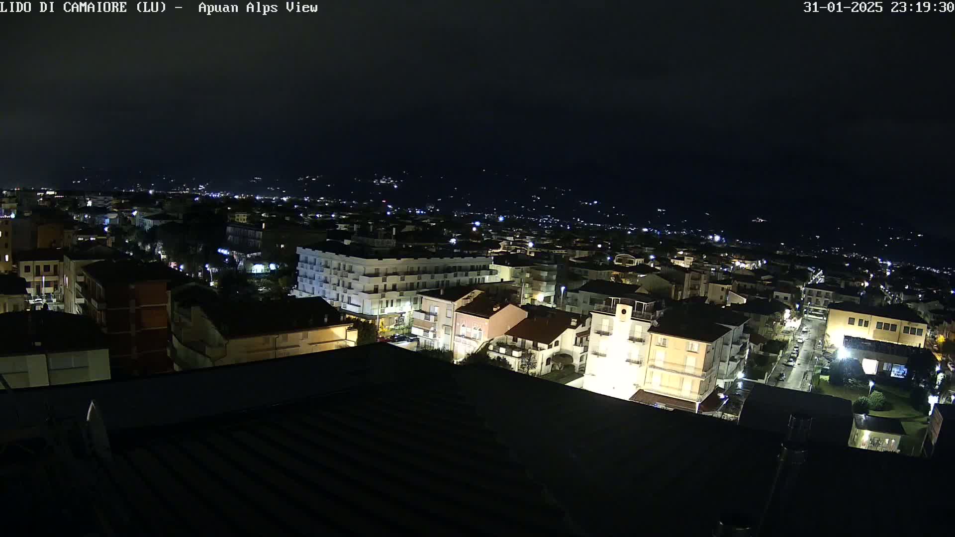A nighttime, high-angle view of a town with illuminated buildings, streets, and distant lights, under a dark sky.