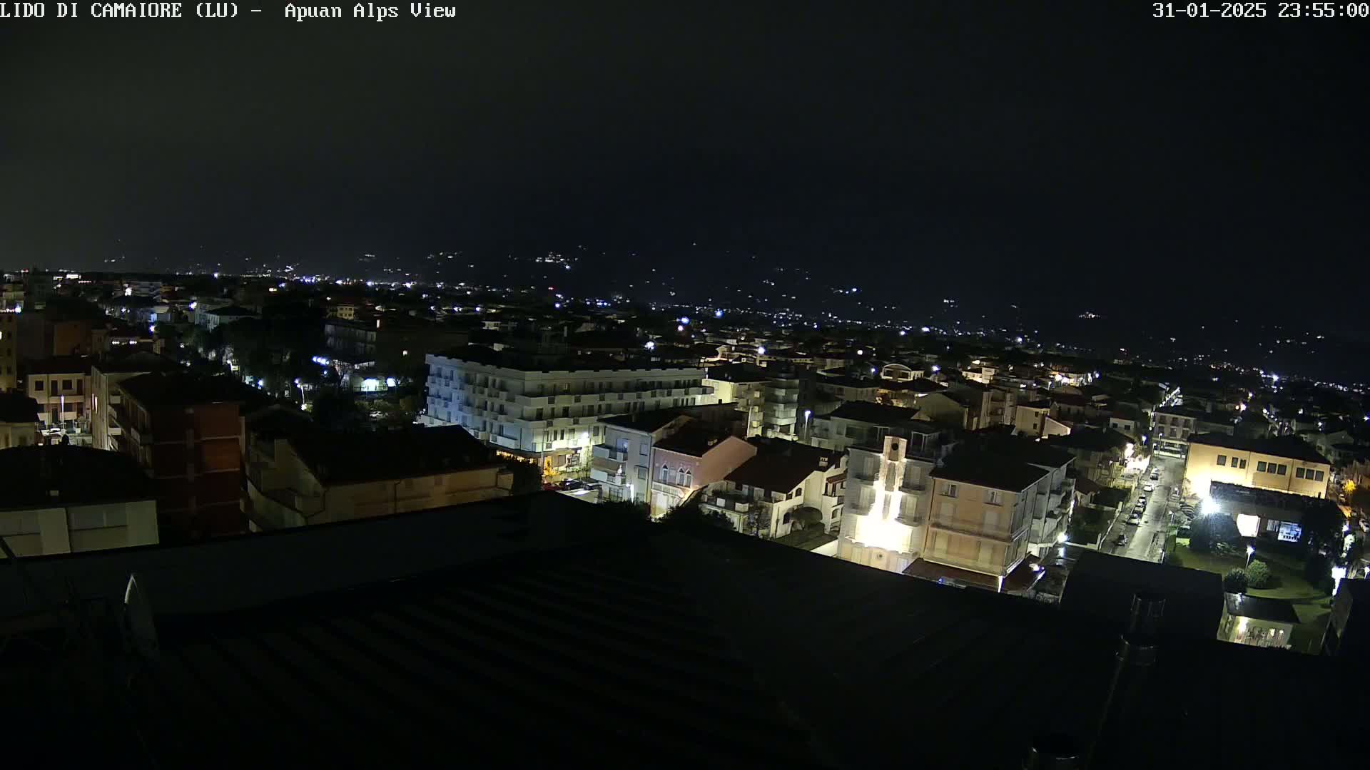 A nighttime aerial view of a town, illuminated by streetlights, with a darker, mountainous background under a clear, dark sky.