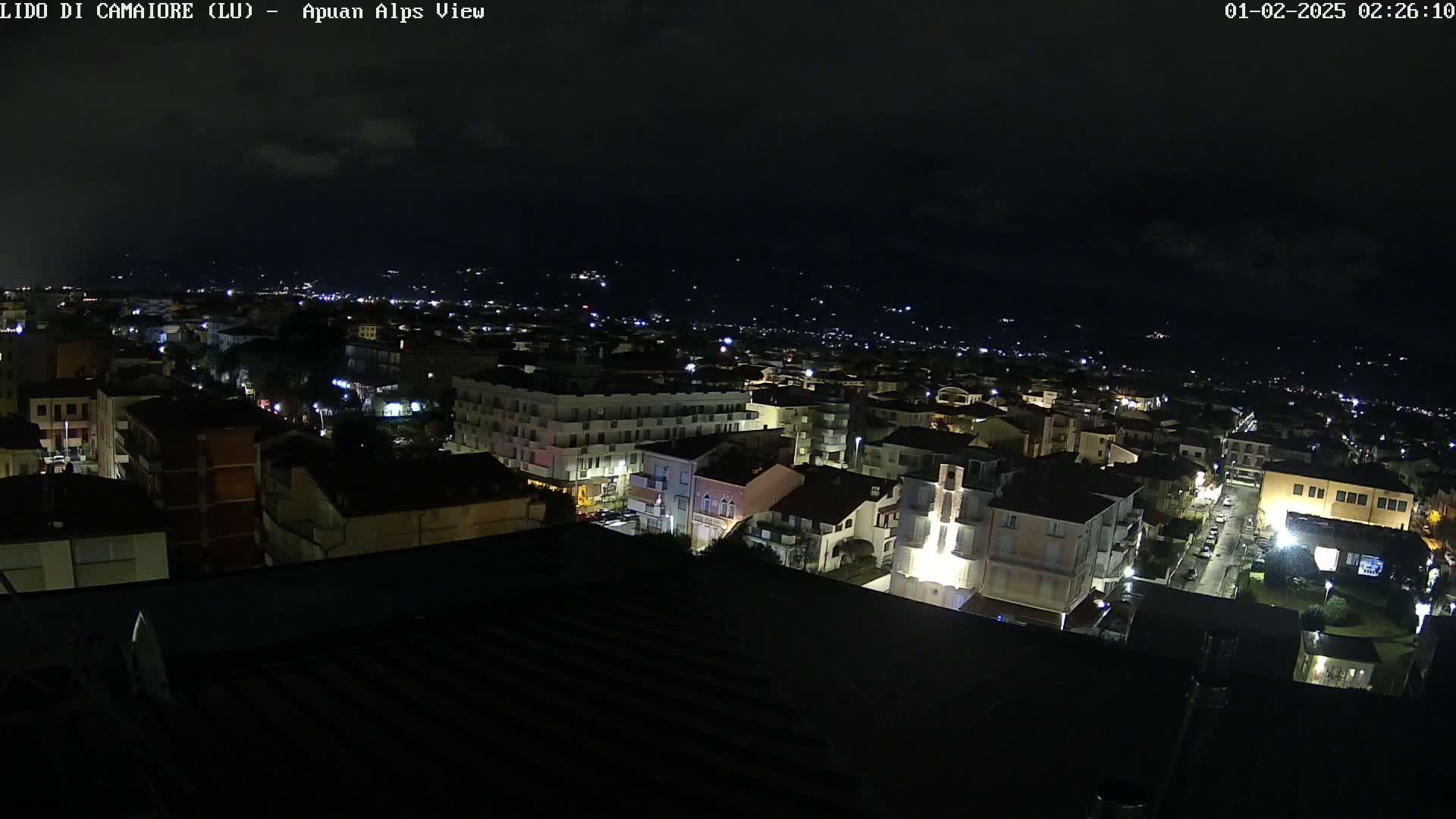 A nighttime, high-angle view of a town with buildings illuminated, extending to a dark mountain range in the distance under a mostly clear sky.