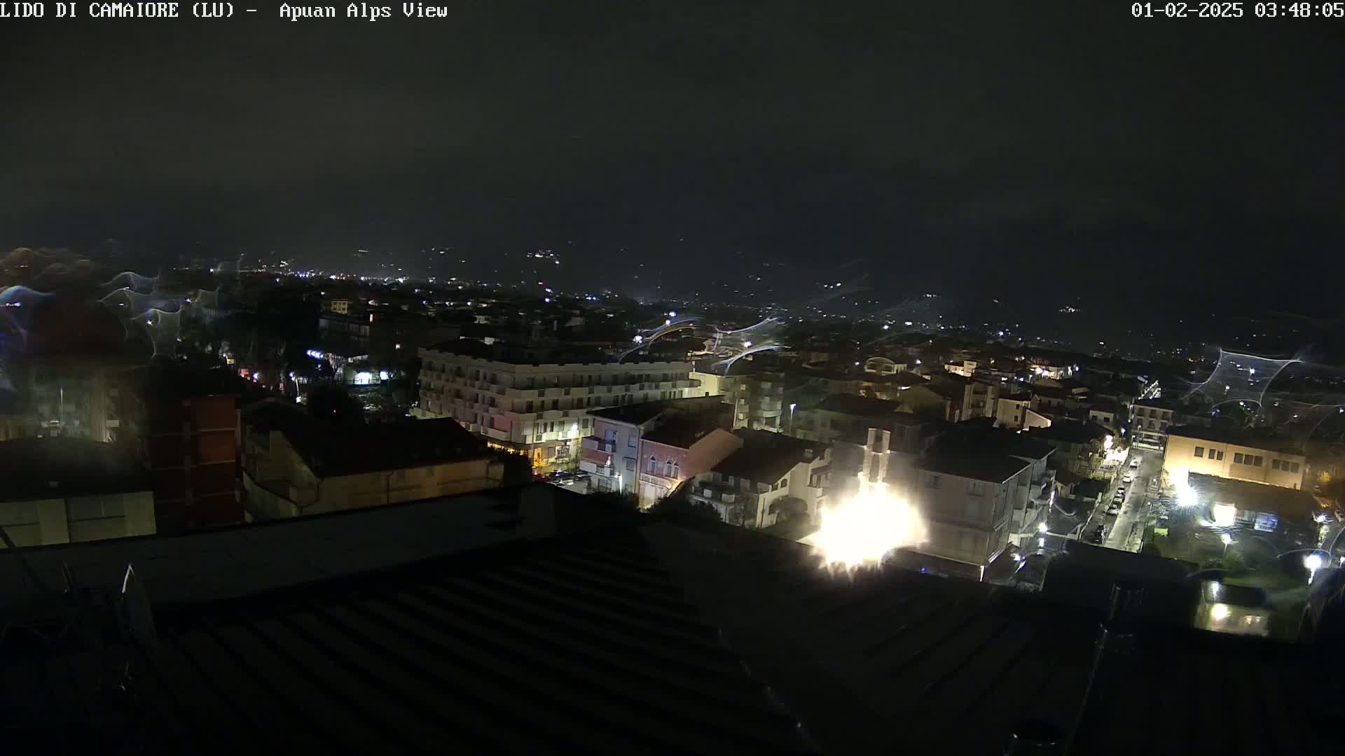 A nighttime, high-angle view of a city illuminated by streetlights and building lights, under a dark sky with streaks of light possibly from rain or wind.