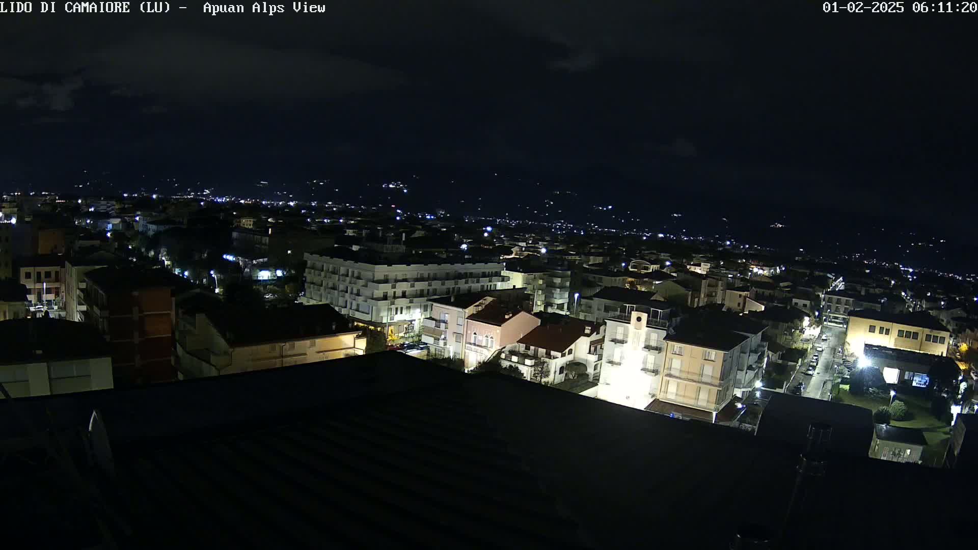 A nighttime, high-angle view of a town illuminated by streetlights, with darker, mountainous terrain visible in the distance under a cloudy sky.