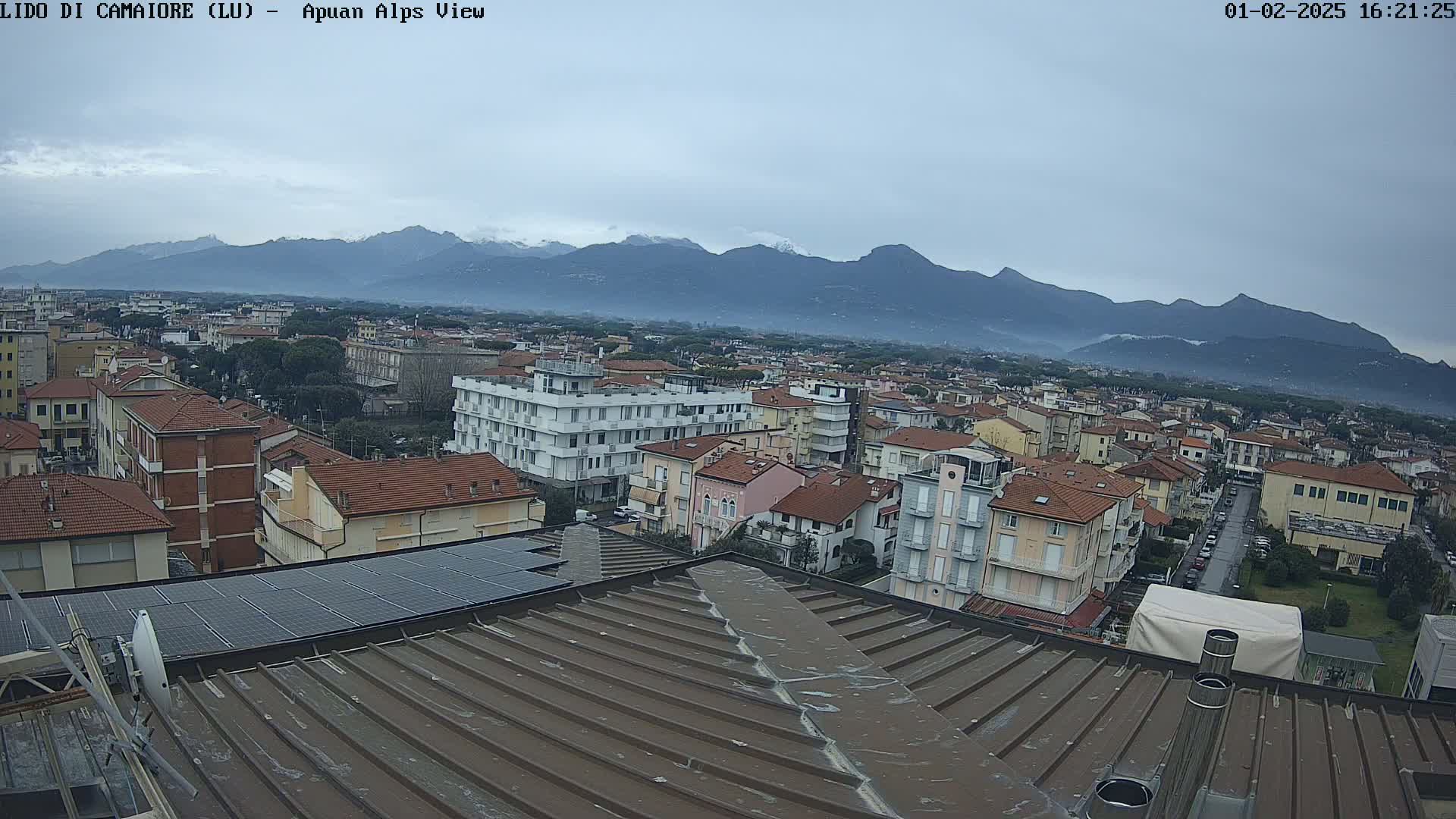 A view from a rooftop shows a town stretching towards snow-capped mountains under an overcast sky.