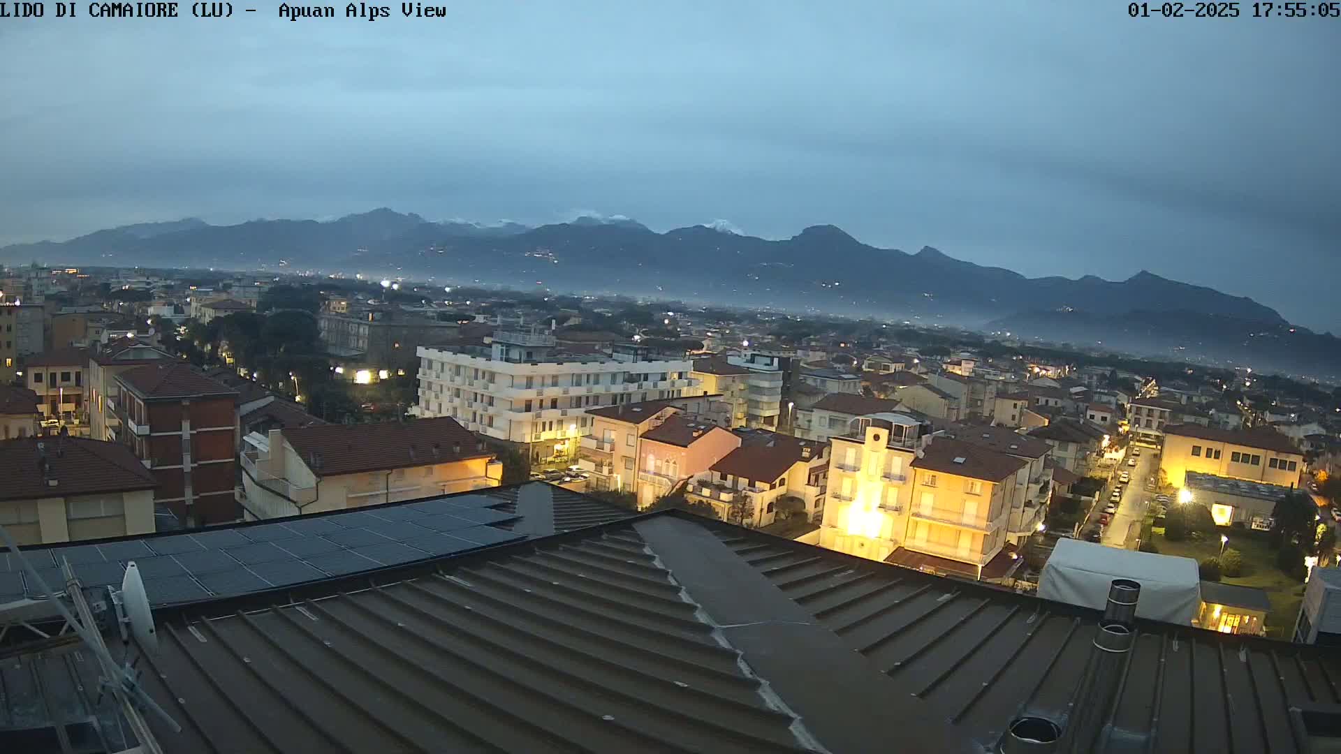 A nighttime view of a town nestled at the foot of snow-capped mountains under a dusky sky.
