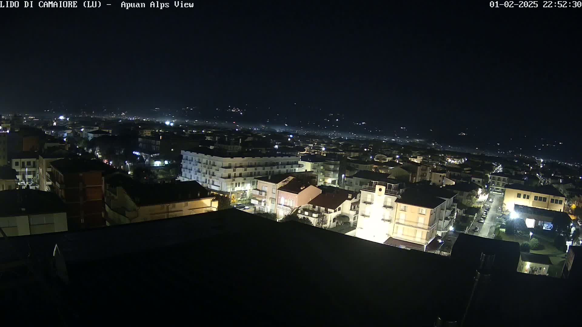 A nighttime aerial view of a town illuminated by streetlights, with faintly visible hills in the distance under a clear, dark sky.